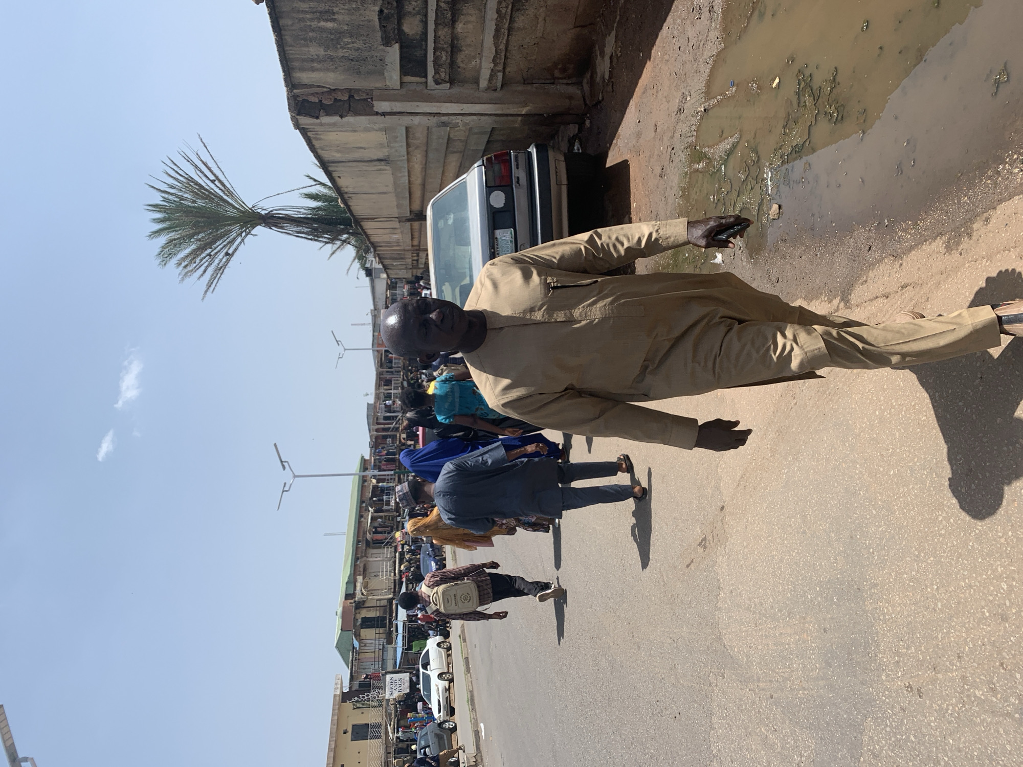 A man in a light brown traditional outfit is walking towards the camera on a paved street in Jos, Nigeria. He has a bald head and is looking down. The street is lined with buildings and market stalls on the left, and a concrete wall on the right. Several other people are walking in the background, some dressed in traditional attire. A silver car is parked on the right side of the street. The weather appears to be sunny and clear. A palm tree is visible above the wall on the right. The ground is uneven and has puddles of murky water on the right side.