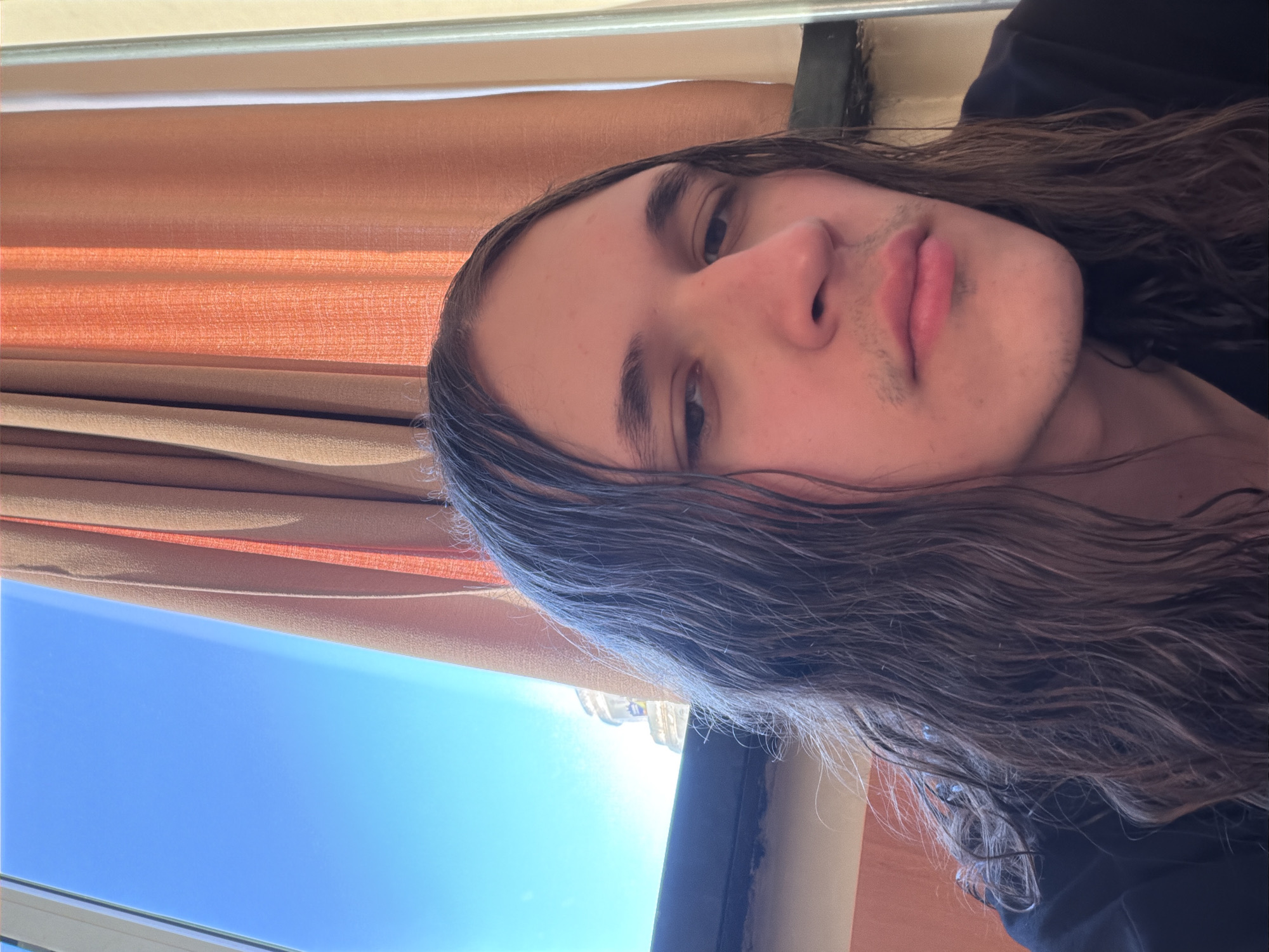The image is a close-up selfie of a young person, likely male, with long, wavy dark brown hair, fair skin, and light stubble on their upper lip and chin. Their head is tilted, and they look towards the camera with a neutral expression, lips slightly pursed. They are wearing a dark-colored top, possibly a t-shirt or hoodie.
The background is split: to one side, there are horizontal orange-brown blinds or curtains, suggesting an indoor or sheltered area. To the other side, a clear, vibrant blue sky is visible, indicating bright, sunny weather. A distant building or structure is faintly discernible against the sunlight, suggesting an urban environment. The sunlight is intense, creating a bright glow in the sky portion of the image.
The photo appears to be taken outdoors or from a window/balcony in Plovdiv, Bulgaria, during daytime on a clear, sunny day. No text is visible in the image.
