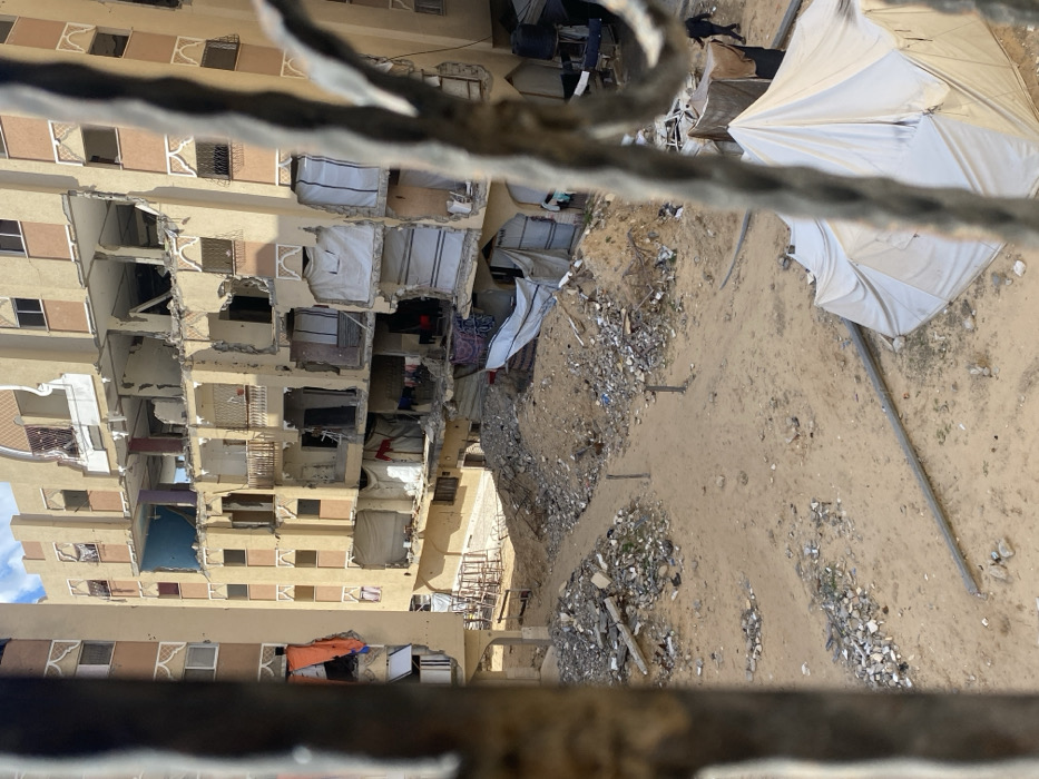 The image captures a severely damaged urban environment in Khan Yunis, Palestinian Territory, viewed through a blurred, dark metal obstruction in the immediate foreground. A multi-story, sand-colored residential building occupies the mid-ground and background, displaying extensive structural damage with multiple floors exhibiting blown-out walls, collapsed sections, and exposed interiors. Numerous windows are empty frames, and some open areas are covered with white tarps or fabric, with laundry visible hanging from lines. To the left, a similar but less visibly damaged multi-story building is partially visible. The ground level consists of sandy, disturbed earth with scattered and mounded piles of concrete rubble and debris. Dirt paths traverse the uneven terrain. In the lower right, a large white tent provides shelter, with a smaller, darker makeshift tent behind it. A long, grey pipe or beam lies horizontally on the sandy ground in front of the white tent. One individual, dressed in dark clothing, is partially visible walking towards the right edge of the frame, near the tents. A stack of chairs is visible on the ground in the mid-left. The sky above is partially cloudy.