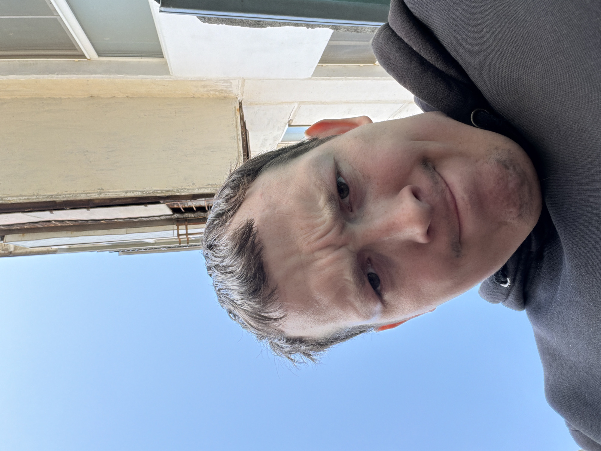 A middle-aged Caucasian man wearing a dark gray hooded sweatshirt is visible in the image. The man's head is angled slightly downward with a scowling expression. The background includes a building with light beige exterior paint and window frames, and a clear blue sky. The image suggests an upward-looking perspective. The location is not specified.