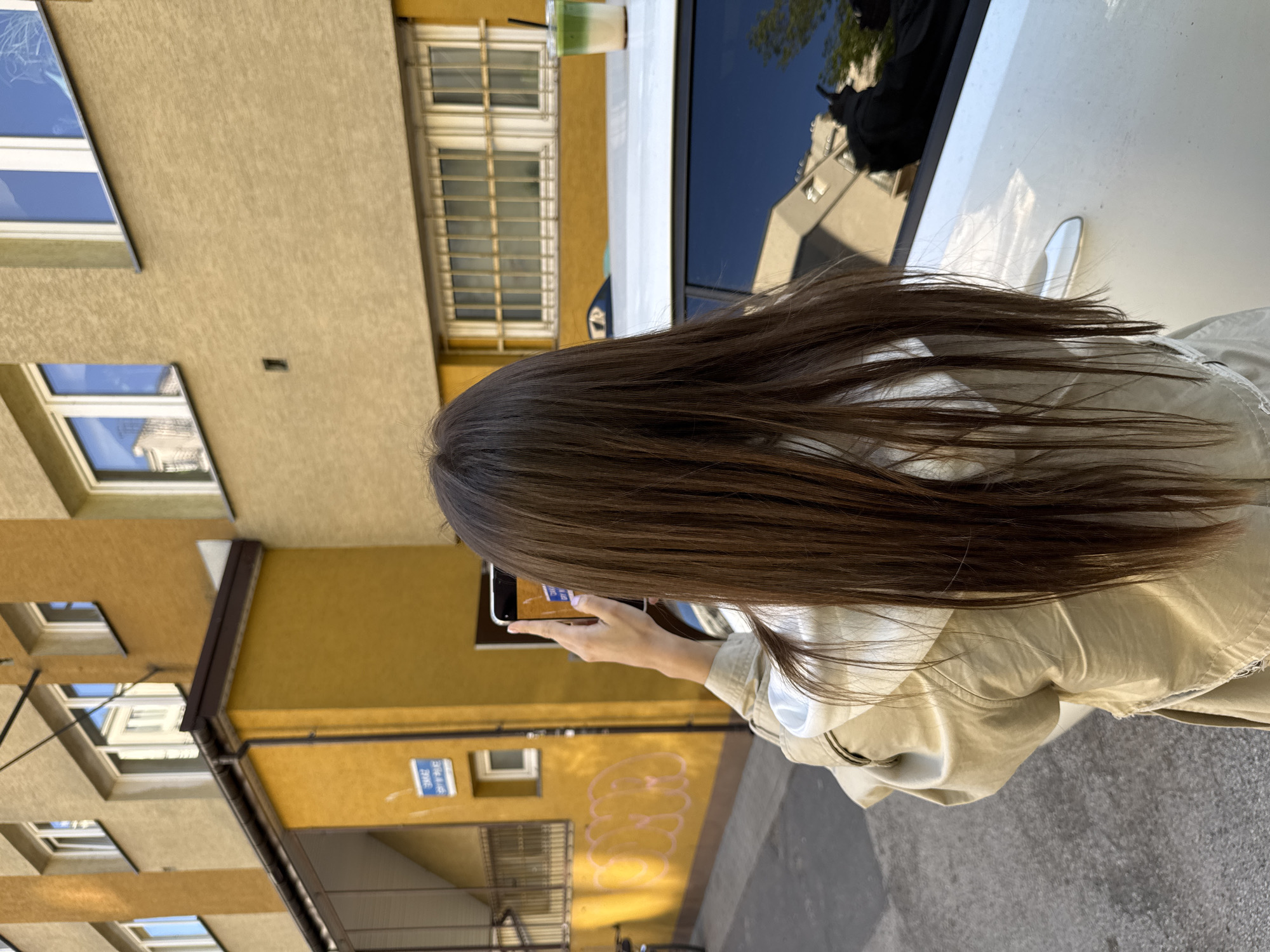 A woman with long brown hair is holding a smartphone in landscape orientation, apparently taking a photo of a building. The building is light yellow and has multiple windows, some of which are reflecting the sky. A blue street sign is visible on the building's facade. There is graffiti near the bottom of the building. Part of a white car, with a beverage on the roof, is also in the image. Based on the architectural style and street sign, the location is likely in Warsaw, Poland. The woman is wearing a light-colored jacket.