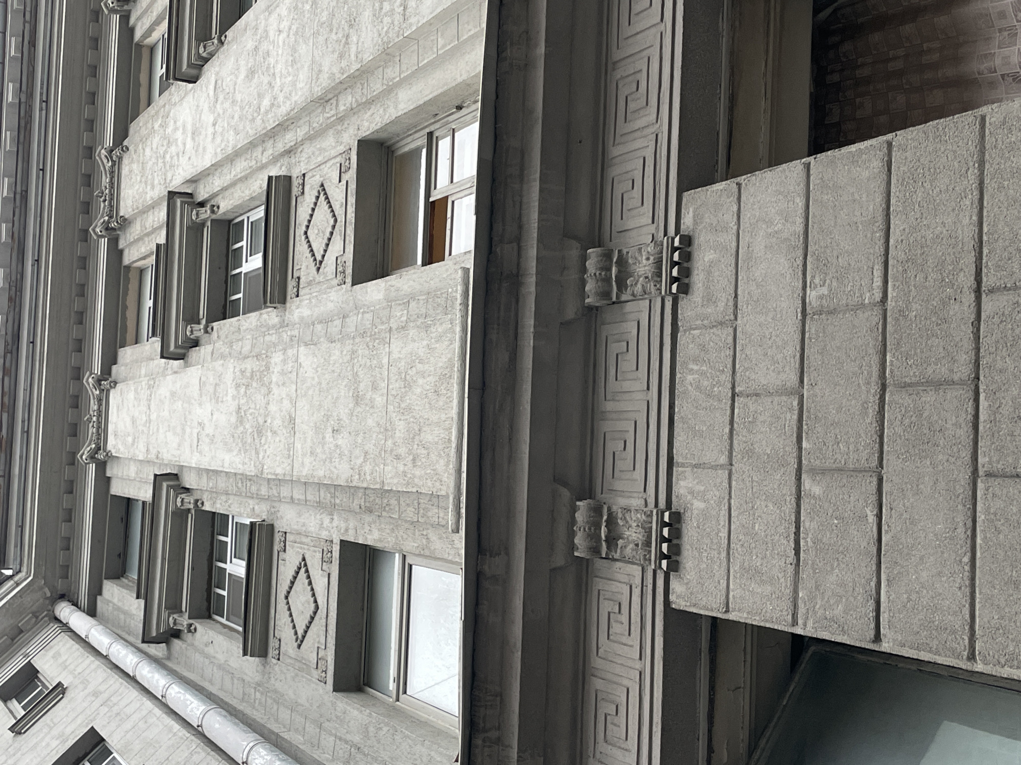 An upward-angled architectural photograph shows a building facade in Saint Petersburg, Russia. The building's exterior is clad in gray stone or concrete, featuring a repeating pattern of windows, each framed with raised stone detailing. Diamond shapes are incorporated into the architectural ornamentation around some windows. A vertical pillar with a Greek key design runs along the right side of the frame. The photograph captures the texture of the stone, revealing a slightly rough, aged surface. A balcony juts out from the building's facade to the right of the pillar. The sky or surrounding buildings are visible through the windows. A drainpipe runs down the side of the building.