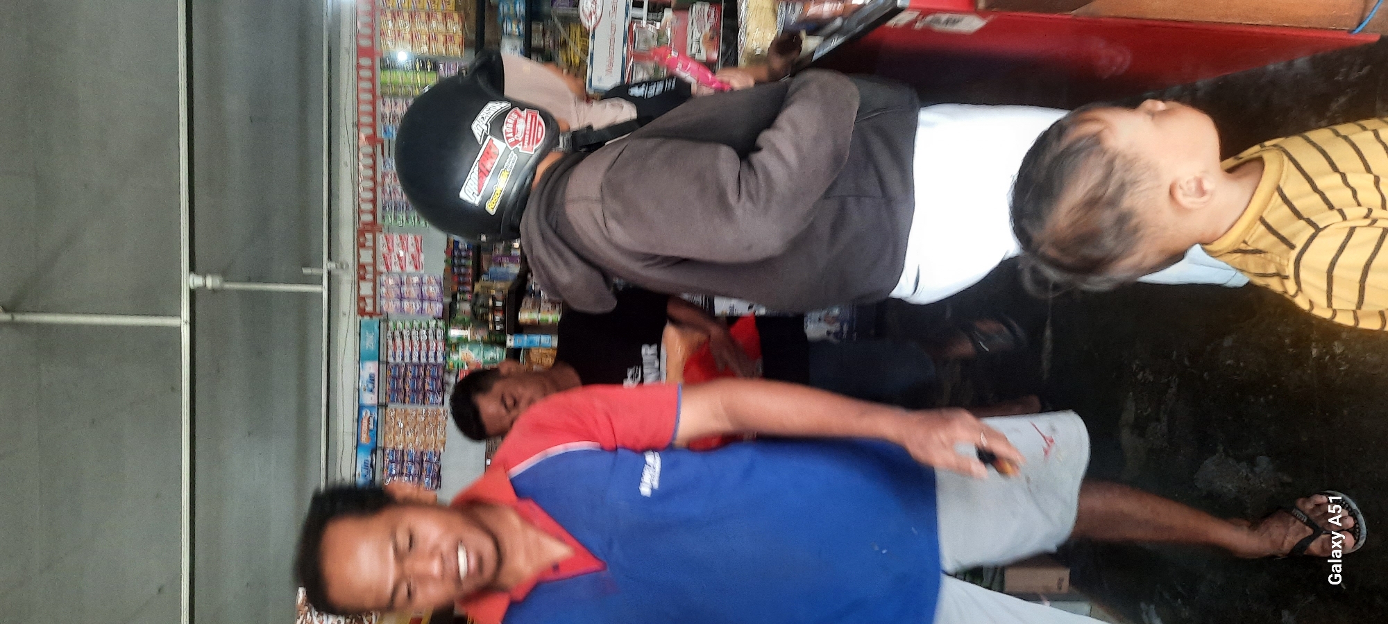 The image depicts an indoor scene, likely a small shop or convenience store, with shelves stocked with various products in the background. In the foreground, a man in a blue and red shirt is partially visible on the left, looking towards the camera with a slight smile. To his right, a man in a black t-shirt is also visible. A person wearing a black helmet with stickers, a brown hooded jacket, and white pants is in the mid-ground, facing away from the camera. A young child in a yellow and black striped shirt is positioned in the lower right of the frame, facing to the right. The floor appears to be dark and wet. A "Galaxy A51" watermark is present in the lower left corner. The setting is likely Selopuro, Indonesia, given the provided context.