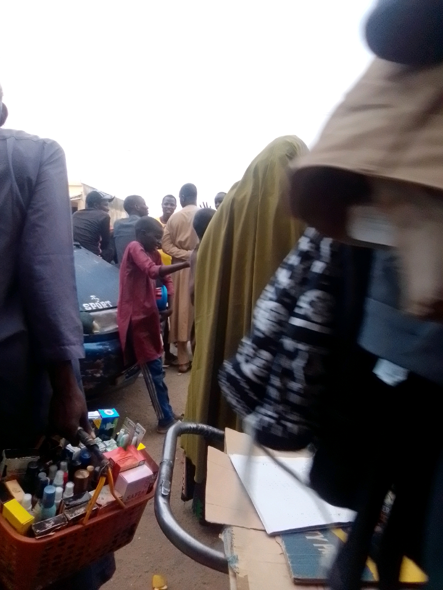 The image depicts a street scene in Arawa, Nigeria. In the foreground, a person with dark skin is holding a basket filled with various small items, including bottles and boxes, possibly for sale. This basket is attached to a metal frame, suggesting it is part of a cart or mobile stall. To the left, a portion of a blue car is visible with the word "SPORT" written on its hood. A young person in a maroon tunic and jeans stands near the car, gesturing with their right hand. In the background, several individuals are gathered, some appearing to be in conversation. The attire of the people, including flowing robes and head coverings, is consistent with the cultural context of Nigeria. The overall environment appears to be an outdoor market or busy street, characterized by an open space with a rough ground surface. The lighting suggests it is daytime, with a bright, possibly overcast sky. The image is taken from a low angle, focusing on the people and the goods being displayed.