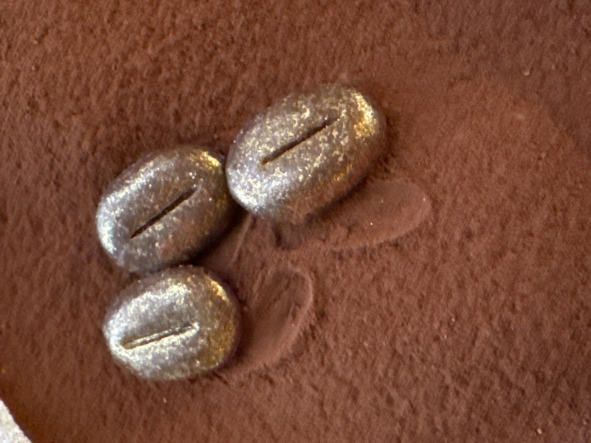 Three ovoid, bean-shaped objects, each approximately 2-3 cm in length, are positioned on a finely textured, reddish-brown powdered surface. Each object is uniformly coated in shimmering, gold-colored glitter and features a distinct longitudinal slit along its top surface, mimicking a coffee bean. They are arranged in a staggered triangular cluster, with two objects in the upper frame and one below and to the right. Faint, shallow indentations are visible in the powder around the objects, suggesting prior contact or movement. The scene is illuminated by direct light, causing prominent specular reflections on the glittered surfaces. The image provides no visual information to determine a specific location within Milan, Italy.