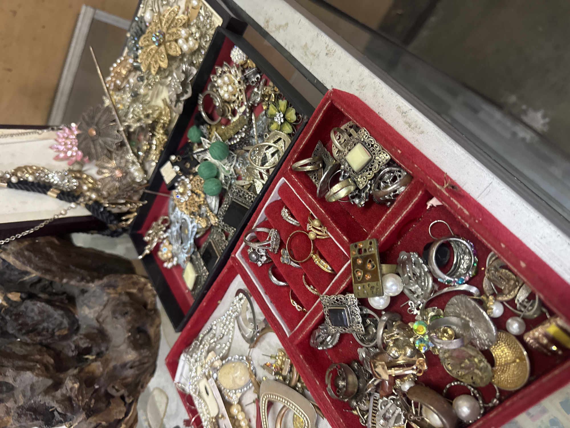 In Denpasar, Indonesia, a close-up shot captures an assortment of vintage jewelry displayed in red velvet trays and a black jewelry box. The jewelry includes necklaces, rings, brooches, and earrings, featuring pearls, gemstones, and ornate metalwork. Some pieces are gold-toned, while others are silver-toned or a mix of both. A rough, dark brown wooden sculpture is visible in the background, adding a rustic element to the scene. The lighting is natural, highlighting the details of the jewelry.