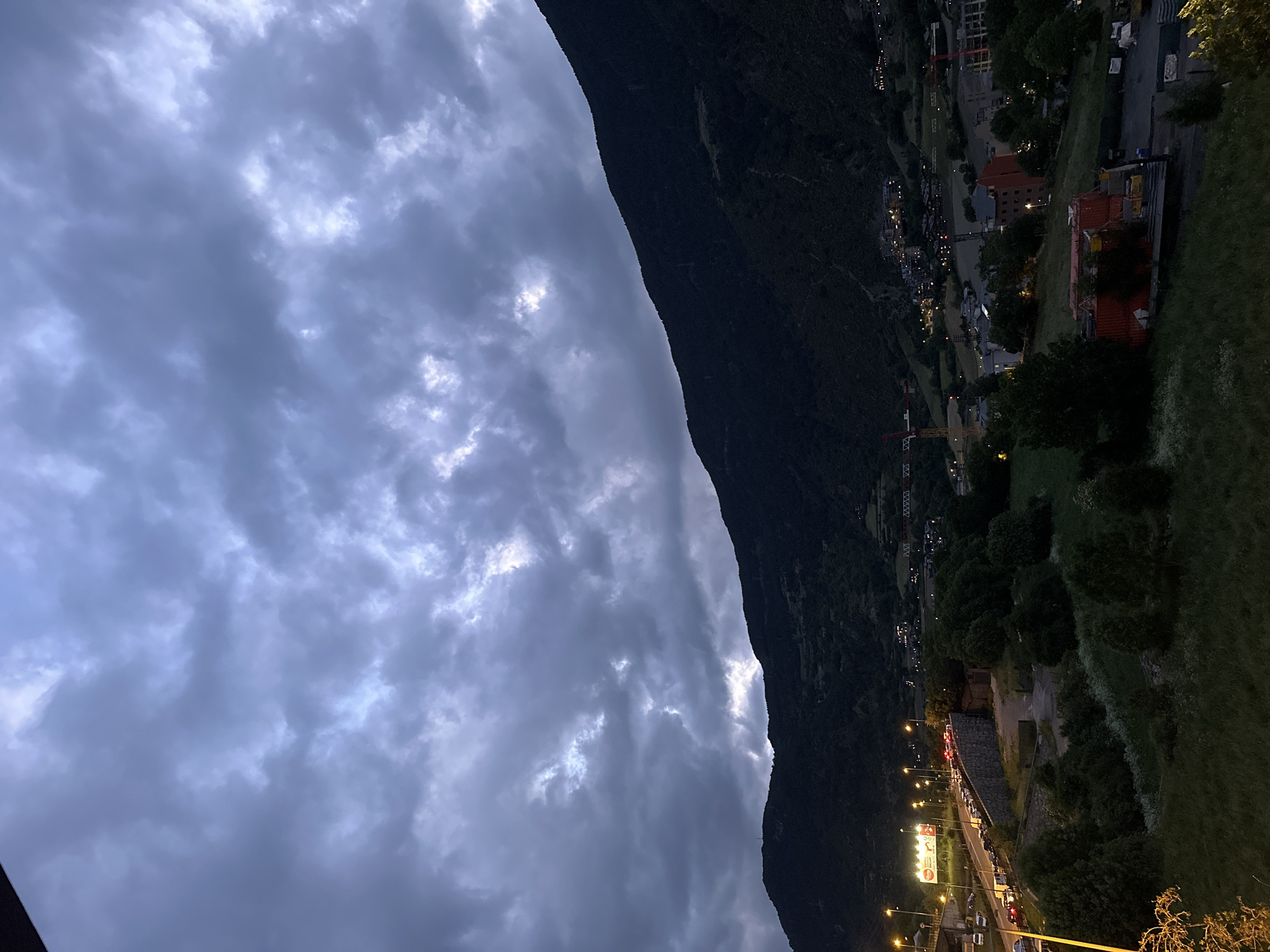 The image presents a nocturnal or dusk scene of a mountainous valley, likely Encamp, Andorra, from an elevated perspective. The sky, occupying the upper left, is heavily overcast with dark, textured clouds, punctuated by lighter patches that suggest reflected light.
A prominent, dark mountain slope dominates the right side of the frame, extending diagonally from the mid-top to the bottom right, showcasing rugged terrain and dense vegetation.
Nestled in the valley below, and stretching across the lower half of the image, is a brightly illuminated town or village. Numerous streetlights and building lights cast a warm glow over the urban landscape. A main road winds through the town, with the streaks of car headlights and taillights indicating active traffic.
Various buildings are visible, some appearing residential, others commercial. Two large construction cranes, one yellow and another white and red, are distinctly visible in the mid-ground, indicating ongoing development within the town. Patches of green foliage and trees are scattered throughout the scene, especially on the mountain slopes and surrounding the settlement. A large, illuminated billboard-like sign stands beside the main road, but any text on it is not clearly legible. The scene conveys a sense of a bustling mountain community against a dramatic, cloudy backdrop after dark.
