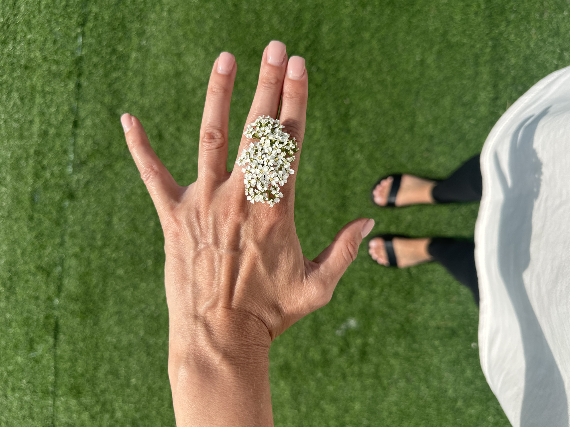 A close-up, top-down shot features a person's hand adorned with a ring made of tiny white flowers. The hand is outstretched with fingers spread, and the ring is positioned on the ring finger. The background consists of green artificial turf, likely a sports field, common in urban recreational areas like those found in Даниловка (Алтайский), Russia. The lower portion of the image shows the person's feet wearing black leggings and sandals, and the edge of a white garment. The lighting suggests it is daytime with bright sunlight.