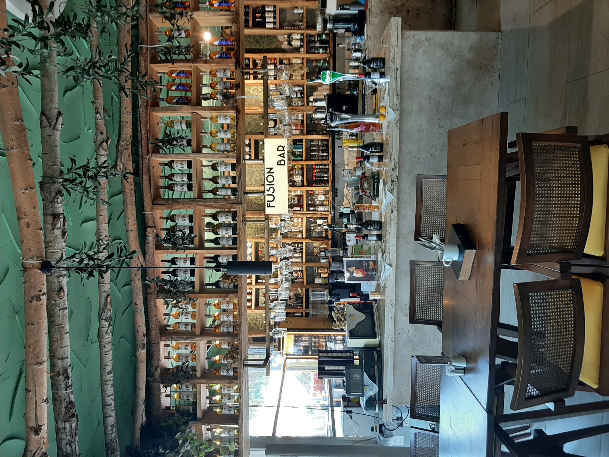 This media file depicts the interior of a restaurant or bar, likely in Alanya, Turkey, as suggested by the context. The scene is dominated by a large wooden shelf unit filled with bottles of wine and other liquors, suggesting a well-stocked establishment. Hanging glasses are also visible on some shelves. In front of the shelving unit is a bar area with various bottles and what appear to be dispensers for drinks. A prominent, illuminated sign reads "FUSION BAR". The overall setting seems to be a daytime scene, with natural light filtering in from a window, though the weather is not discernible. The style of the bar and shelving, along with the presence of tables and chairs, indicates a commercial establishment designed for serving customers. The decor includes decorative tree trunks and a green textured wall on the left, adding a unique aesthetic. The absence of people implies the establishment might be closed or that the photo was taken before opening or after closing. The scene evokes a sense of an inviting and perhaps upscale dining or drinking establishment.