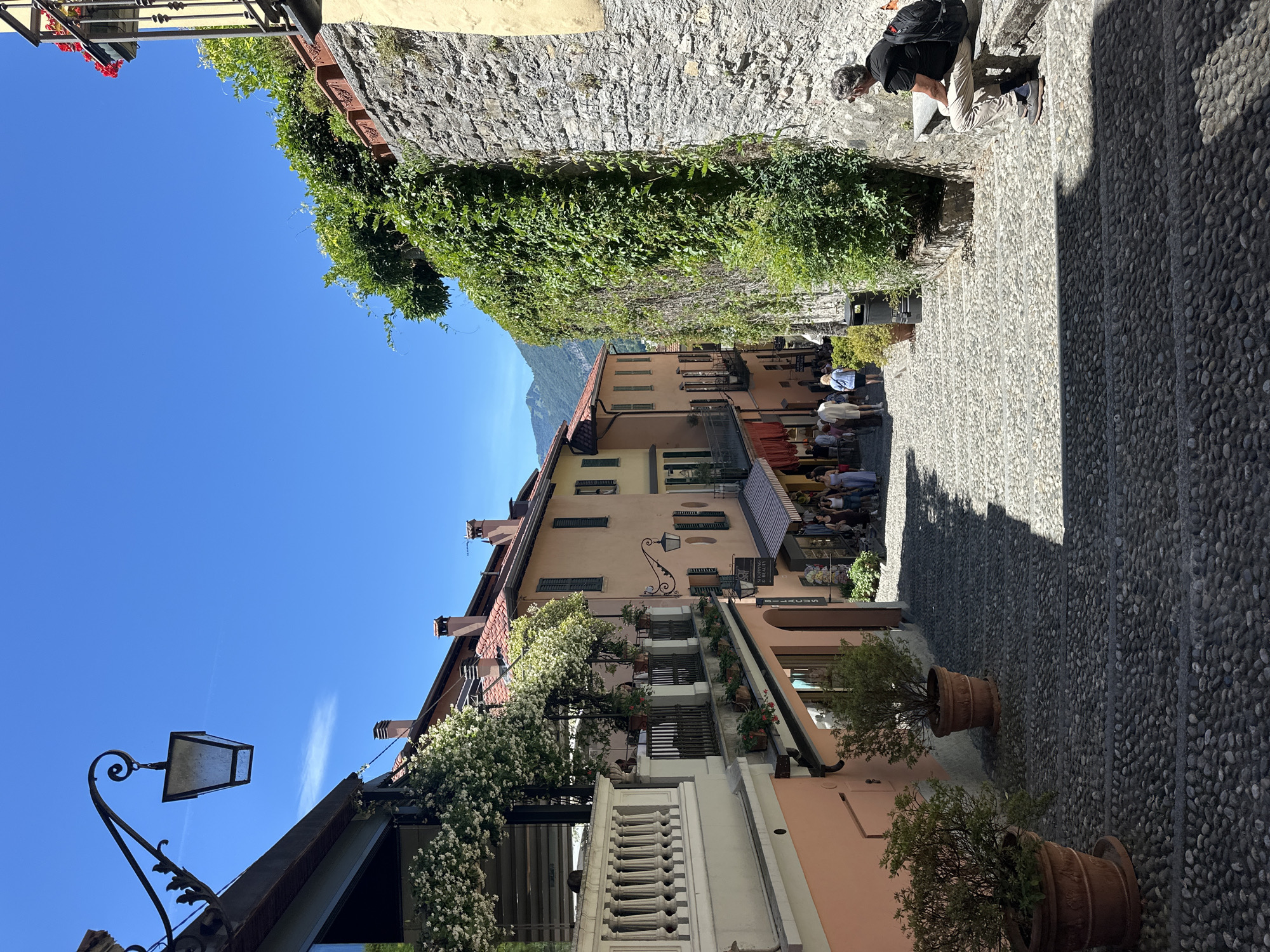 The image depicts a sun-drenched cobblestone street in San Giovanni, Italy, on a clear day. The street slopes downwards and is lined with colorful buildings featuring traditional tiled roofs and shutters. Lush greenery, including flowering vines and potted plants, cascades from balconies and climbs stone walls, adding a vibrant contrast to the architecture.
In the foreground, large terracotta pots with green plants sit on the cobblestone steps, casting sharp shadows. Further down the street, a group of people, appearing to be tourists, walk and browse shops. One individual, a man with graying hair wearing a black t-shirt, beige pants, and a backpack, sits on a stone bench on the right side of the street, looking away from the camera. The sky is a brilliant, cloudless blue, with distant mountains visible in the background, suggesting a picturesque Mediterranean setting. The overall atmosphere is one of a charming, historic European town on a warm summer day.