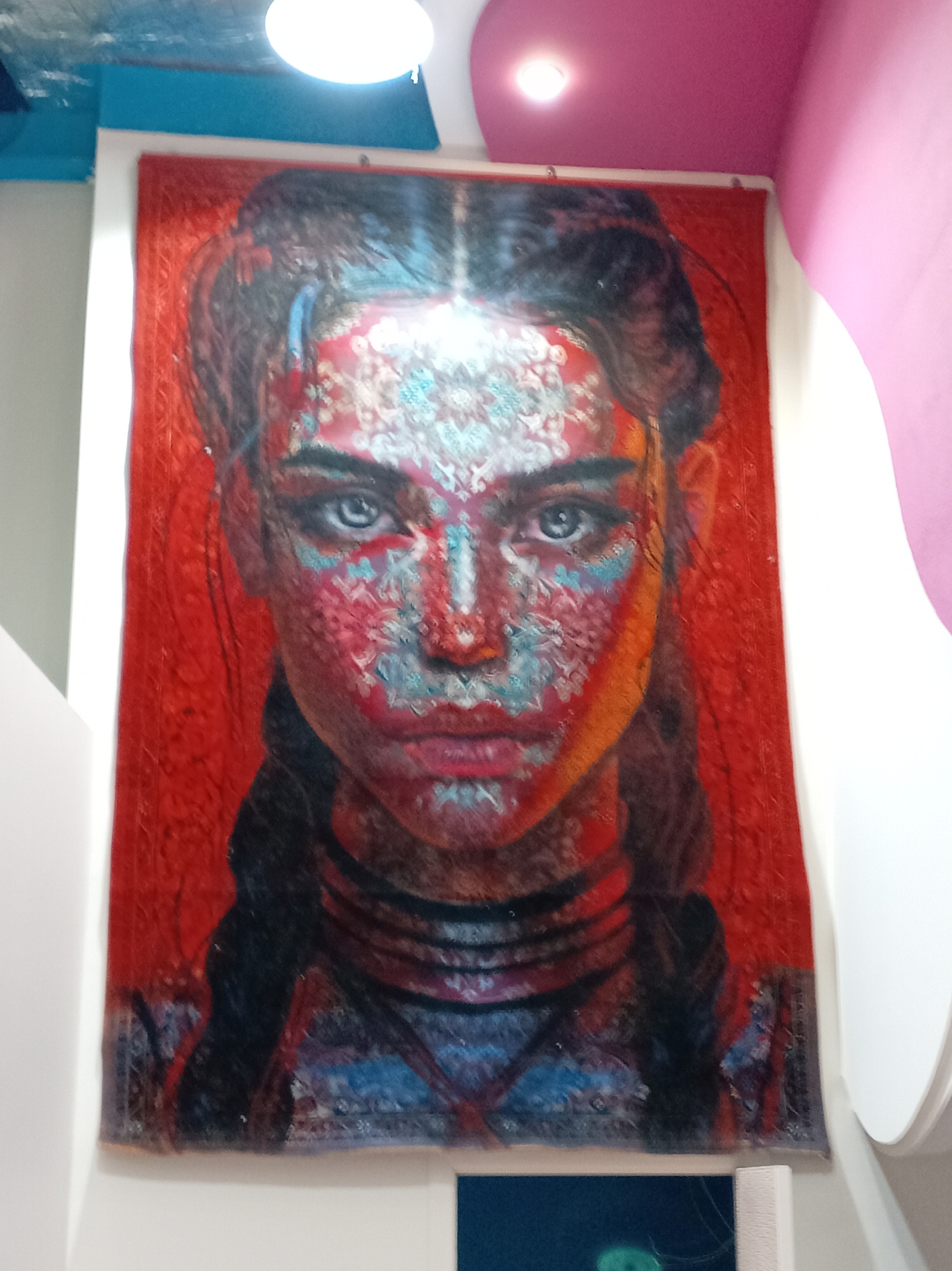 The image shows a portrait hanging on a white wall. The portrait depicts a woman with dark hair in braids. Her face is painted with intricate patterns in shades of red, white, and blue. The background of the portrait is a solid red color. Above the portrait, there's a white and pink ceiling with recessed lighting. The portrait seems to be hanging in a room with modern interior design. The location and the city are unknown.