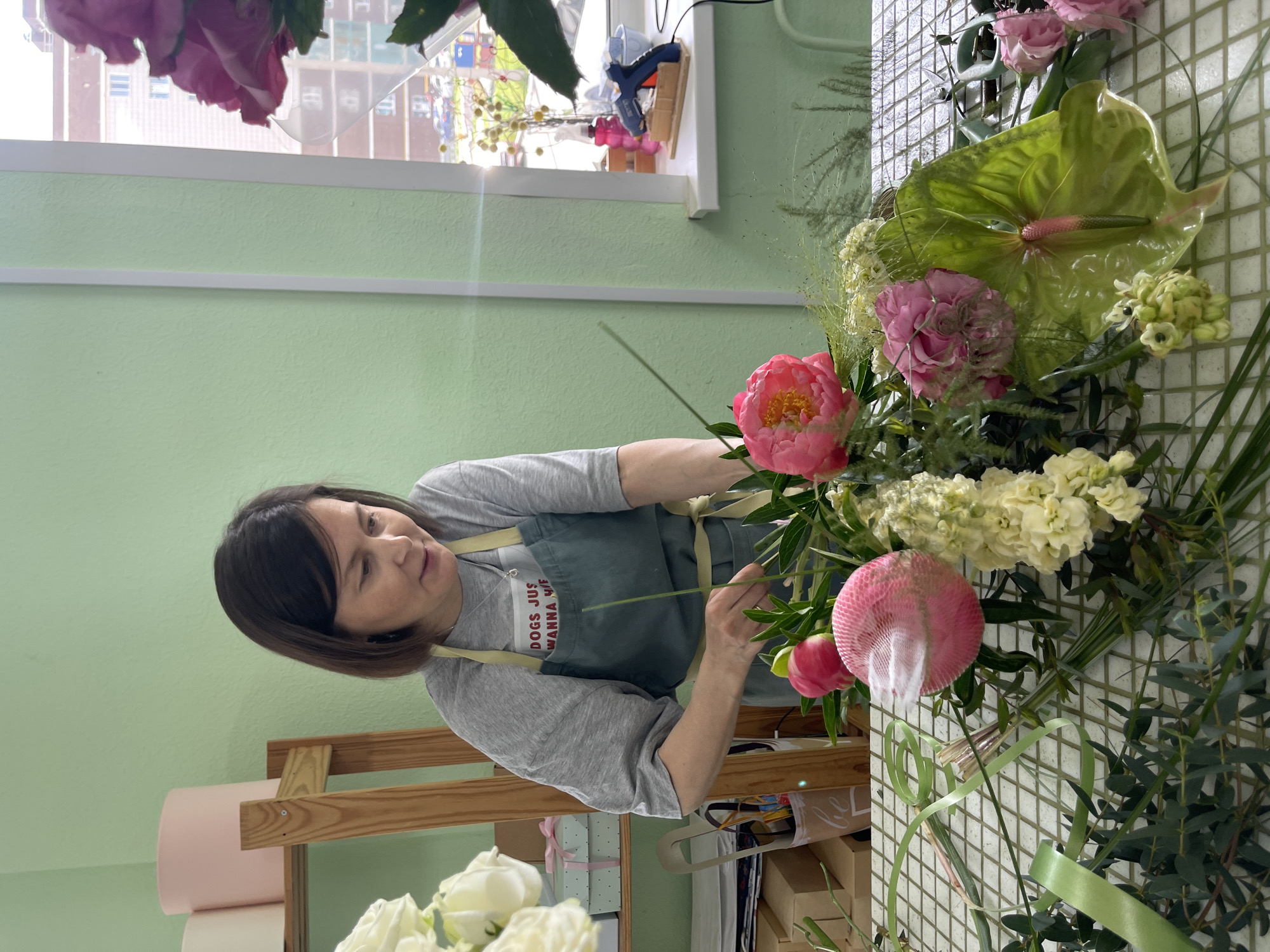 A medium shot captures a woman in a light green apron over a grey t-shirt arranging a vibrant bouquet of flowers. The woman, with dark hair and fair skin, is focused on her task, holding floral stems in her hands. She is positioned in a brightly lit room with mint green walls, suggesting a workshop or floral studio.
The bouquet itself is a rich arrangement of pink peonies, delicate white and yellow filler flowers, and a prominent, large green anthurium. Other floral elements, including eucalyptus branches and wispy green stems, are also visible. The flowers are laid out on a white tiled surface, creating a grid pattern.
On a shelf behind the woman, pink and white cylinders, possibly rolls of floral paper, are stacked. To the right of the scene, a window shows a blurry outdoor view of buildings and what appears to be a playground. On the windowsill, a pink spray bottle, a blue glue gun on a stand, and a small blue bowl are present.
The overall impression is one of a creative workspace where floral arrangements are being meticulously crafted.