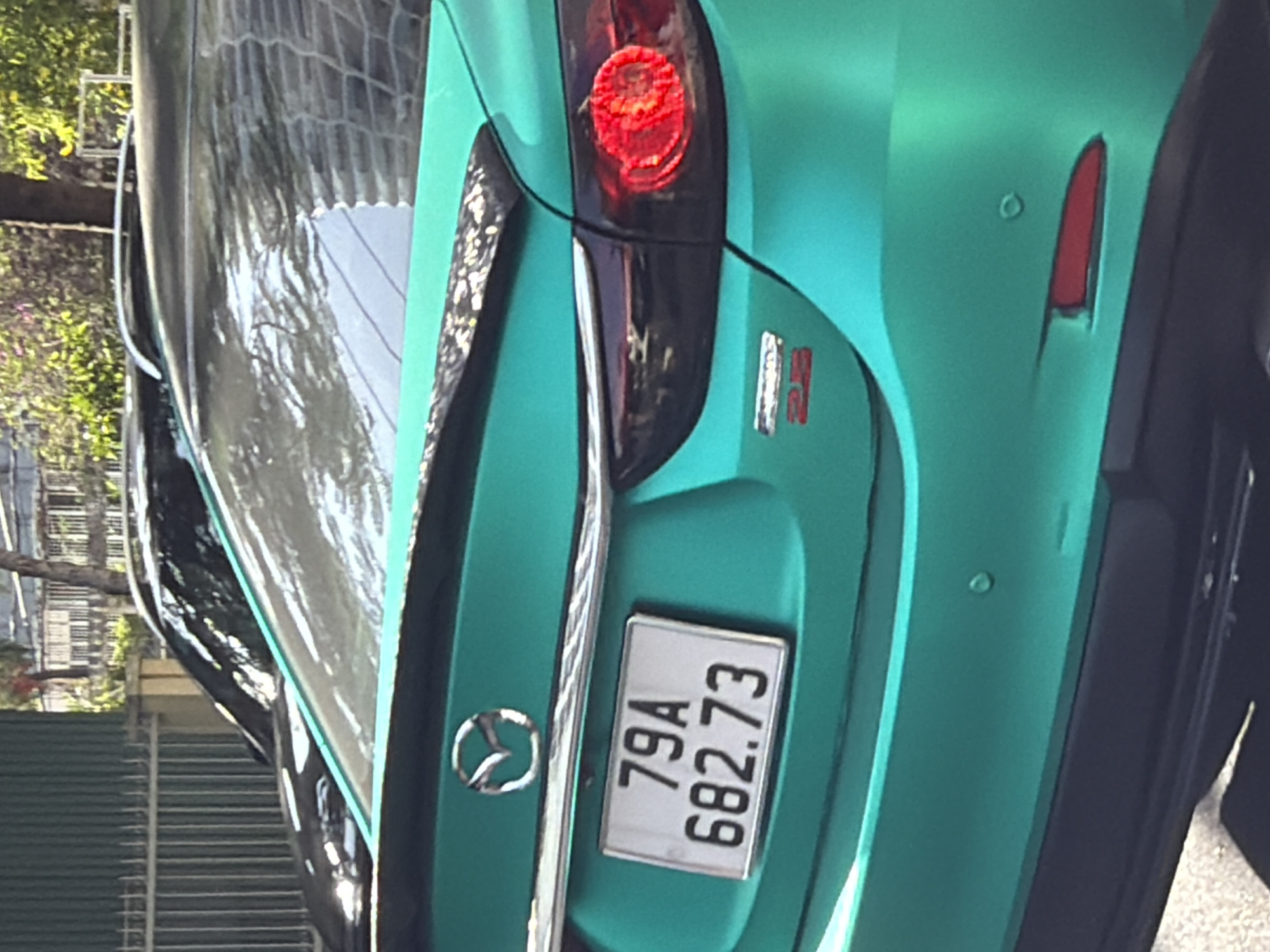 The image shows the rear of a turquoise-colored Mazda sedan, identified by the emblem on the trunk. A license plate with the characters "79A 682.73" is visible. The car is parked on a street, with a metal fence and buildings in the background, suggesting an urban setting. The overall condition of the car appears to be good. The license plate indicates the vehicle is registered in Khanh Hoa province, Vietnam, where Nha Trang is located.