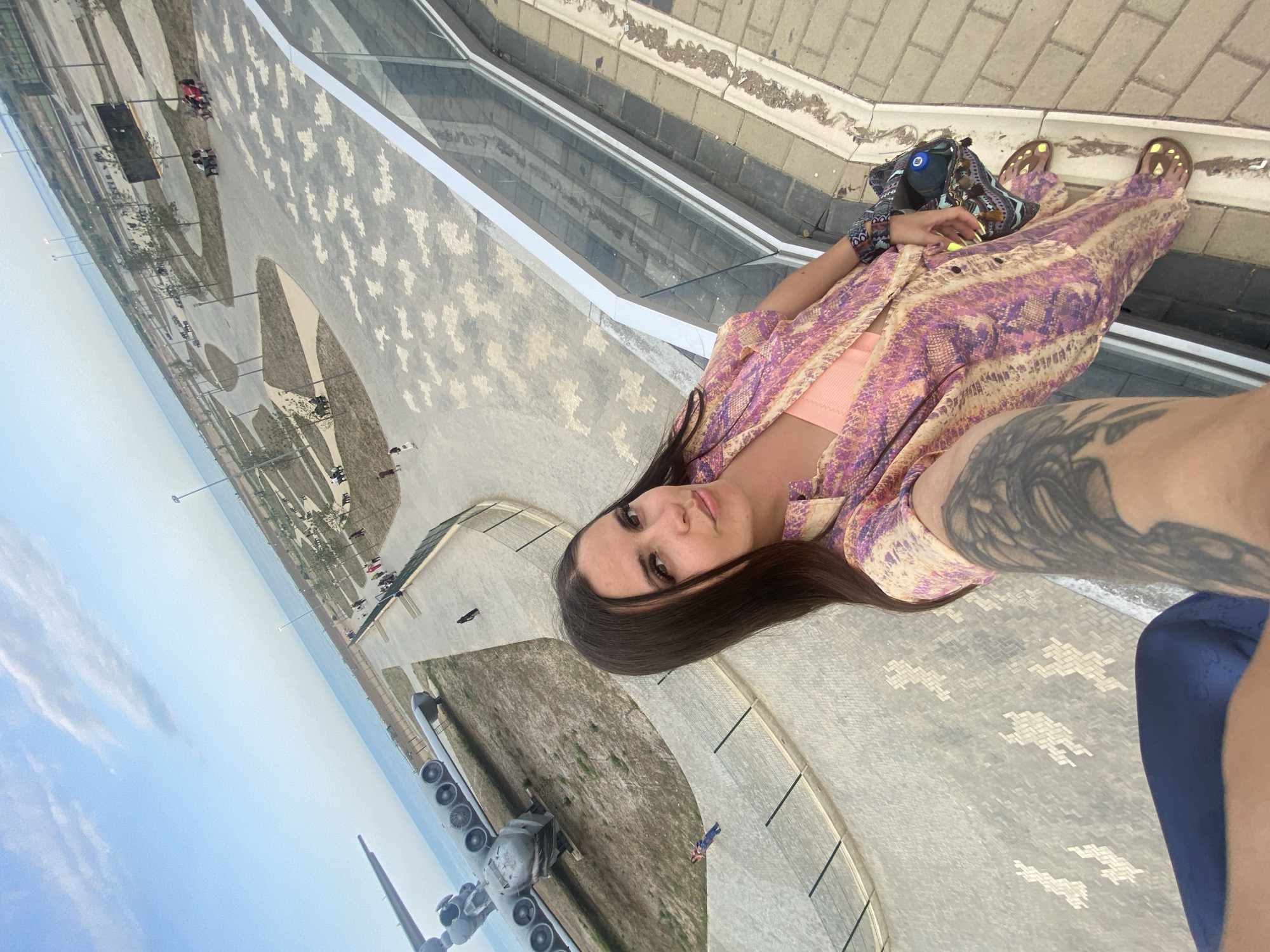 A woman with long, dark hair is taking a selfie at an outdoor park. She is wearing a pink and purple patterned shirt and pants, and an orange top underneath. She has a tattoo on her arm, and is wearing yellow nail polish. In the background, an airplane is on display, and there is a body of water visible. The park has paved walkways, benches, and trees. There are other people in the park, some sitting on benches and others walking. The sky is blue with some clouds. The location is the Arablyar city park in Russia.