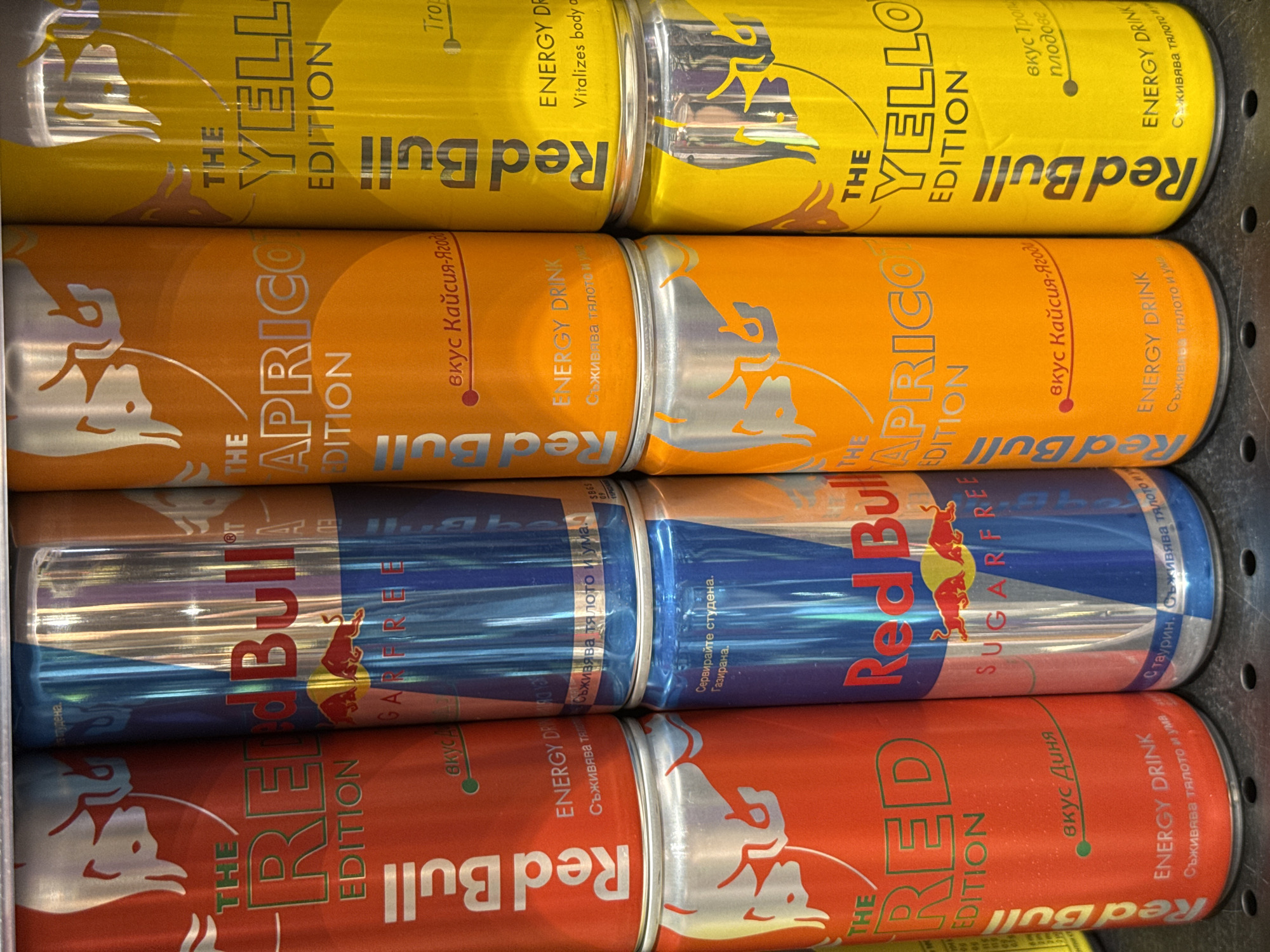 The image shows a close-up, eye-level view of several stacked cans of Red Bull energy drink in a retail setting, likely a supermarket or convenience store. The cans are arranged in rows, with the front row containing four cans: a red one on the far left labeled "RED EDITION", a blue one next to it labeled "SUGARFREE", an orange one labeled "APRICOT EDITION", and a yellow one on the far right labeled "YELLOW EDITION". The cans behind these are partially visible, showing similar Red Bull branding and colors. The lighting is bright and even, typical of an indoor retail environment. The background is blurred, suggesting a shallow depth of field. No people are visible in the image. This photograph was taken in Pleven, Bulgaria.