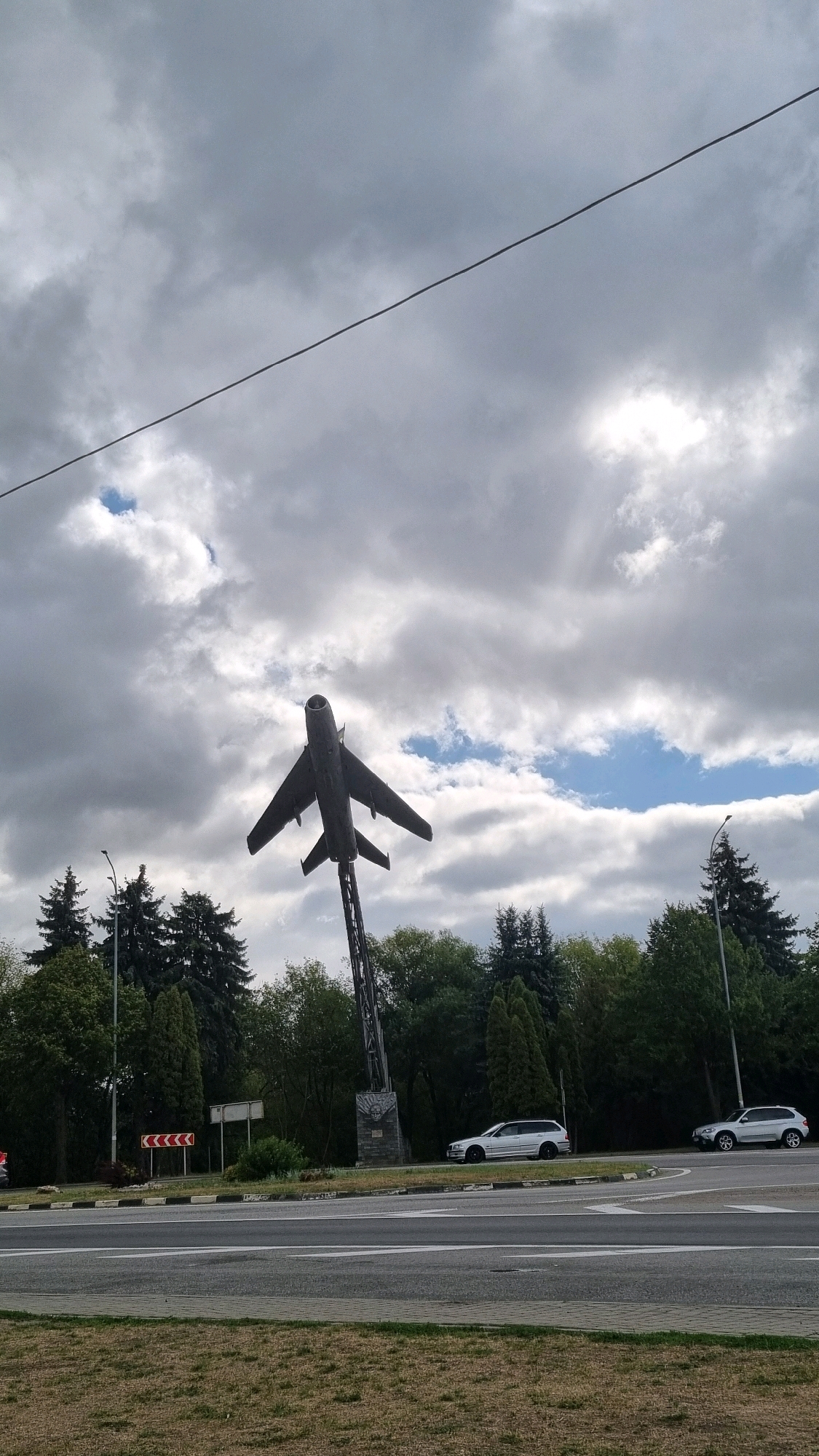 A monument of a military jet is displayed on a tall metal pole. The jet is dark gray and appears to be a Soviet-era fighter jet. It is mounted in a dynamic pose, as if in flight. The monument is located at a traffic intersection in Dubno, Ukraine. Several cars are driving on the road around the monument. The sky is cloudy, with patches of blue visible. The trees surrounding the intersection are lush and green, suggesting it is either spring or summer. The overall scene is a common sight in many post-Soviet cities, where monuments to military history are often incorporated into public spaces.