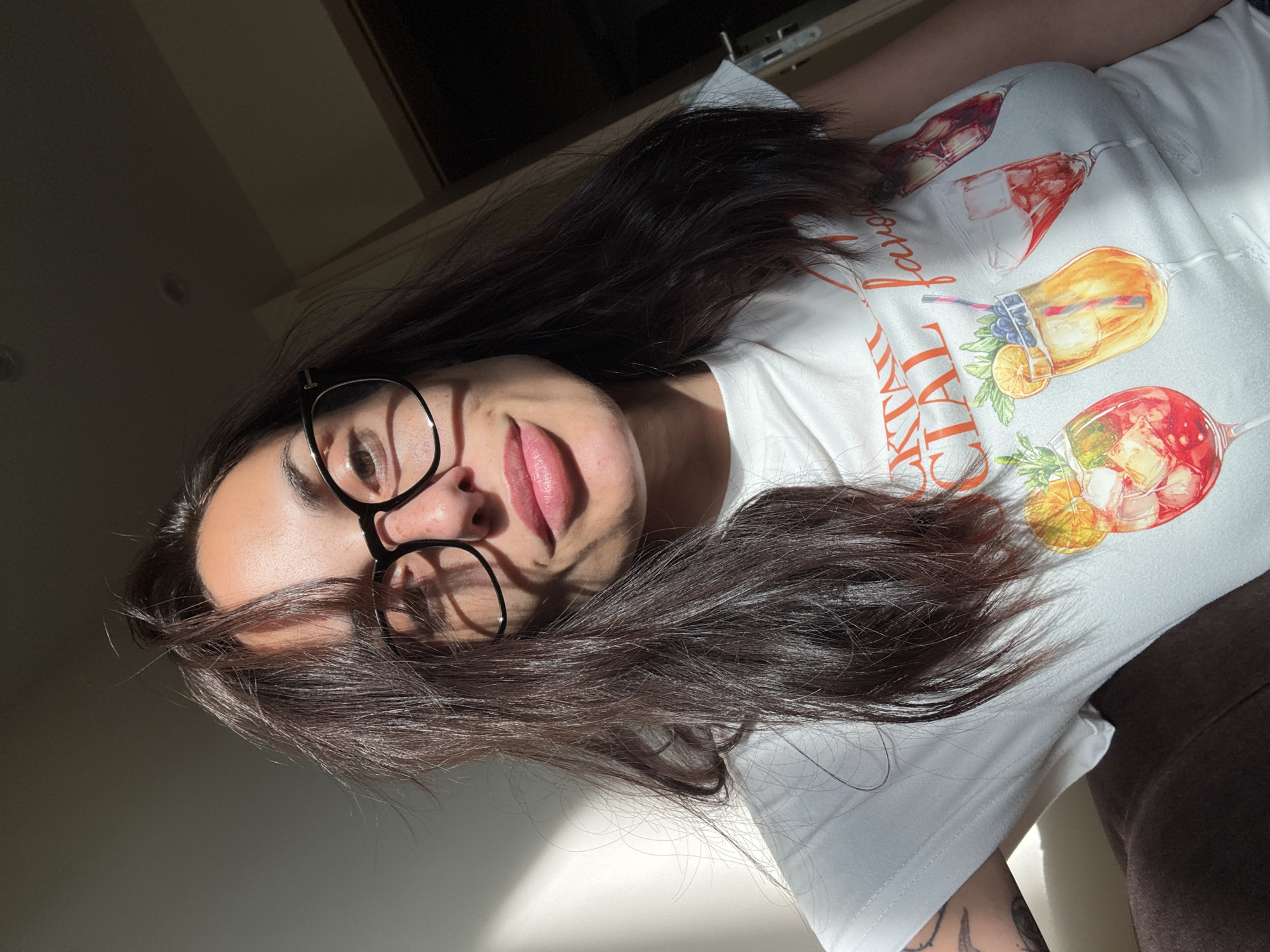A woman with long dark hair, wearing black-rimmed glasses and a white t-shirt with cocktail images printed on it, is visible. Part of her tattooed arm is visible. The background appears to be an interior with sunlight streaming in. There is no evidence to suggest this image was taken in Pleven, Bulgaria.