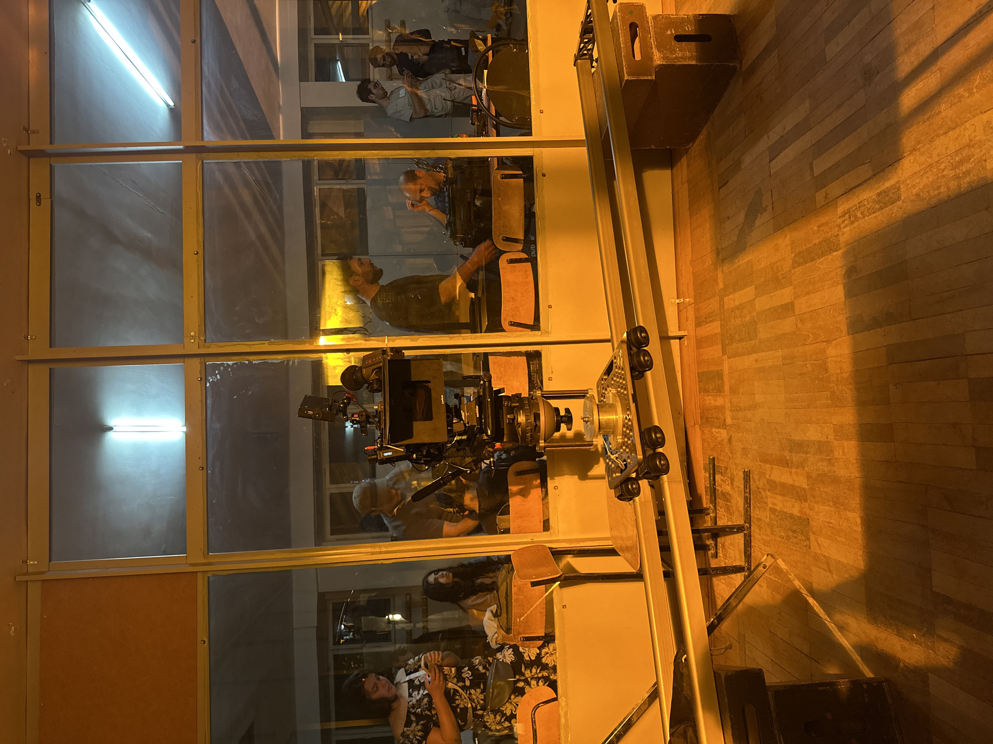 The image shows a film set, likely indoors, in Yerevan, Armenia. A large film camera is mounted on a dolly, positioned near a wall with multiple window-like sections. Reflections in the glass reveal several people, including actors and crew members in different poses and engaging in various activities, suggesting a scene being filmed. The floor is made of light brown wooden planks. There is a shadow on the right side, potentially cast by a stand. The presence of filming equipment and individuals in different states of action indicates an active movie or television production.