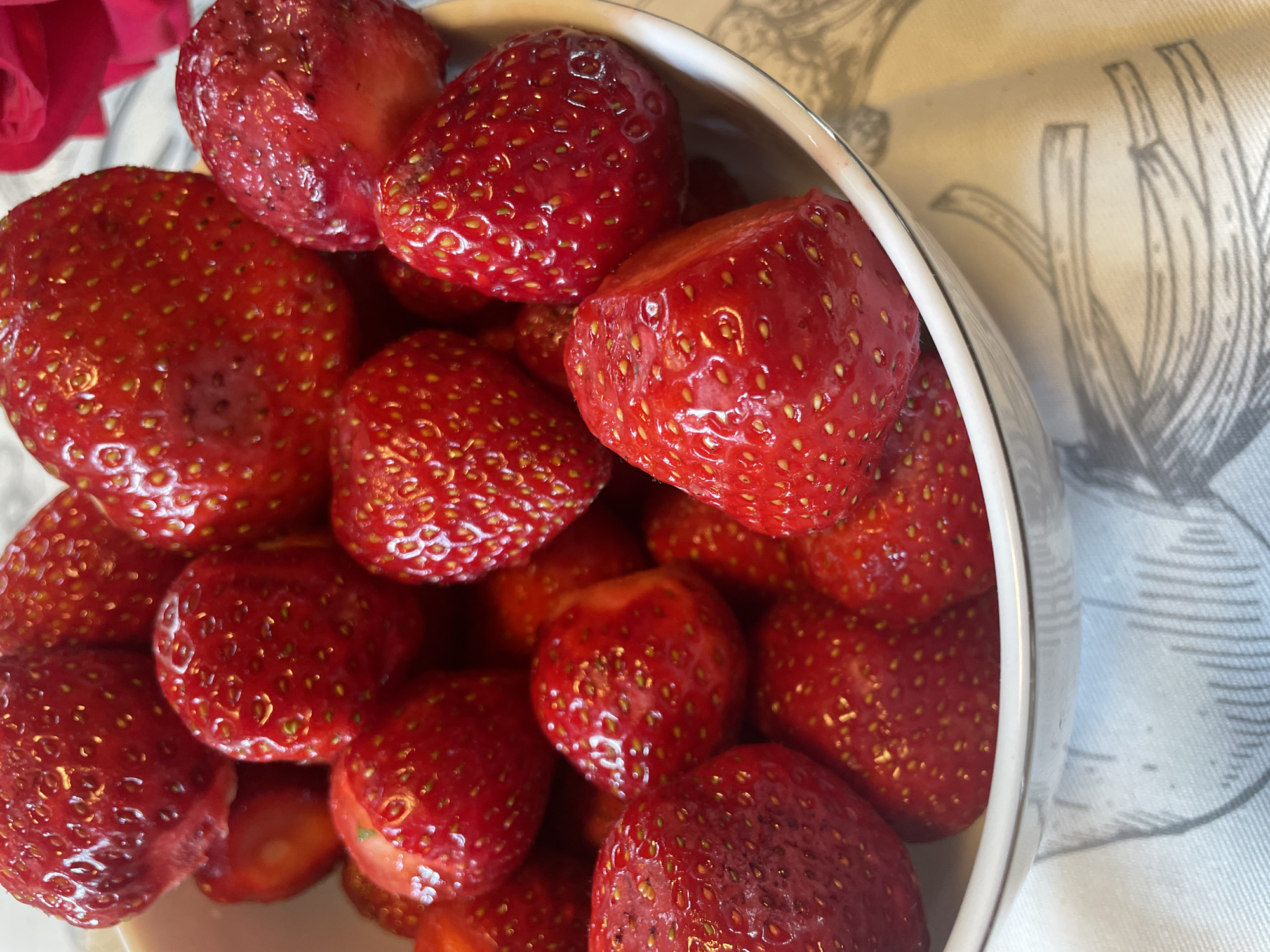 A close-up, slightly elevated shot captures a white bowl filled with plump, ripe strawberries. The strawberries are a vibrant red, with visible seeds dotting their surfaces. Some strawberries have a dewy sheen, suggesting they are freshly washed or have a natural glaze. The bowl is positioned on a white cloth with a faint gray floral or botanical pattern. A blurred pink or red rose petal is visible in the upper right corner of the image. The lighting highlights the texture and juiciness of the fruit. The location is not specified.