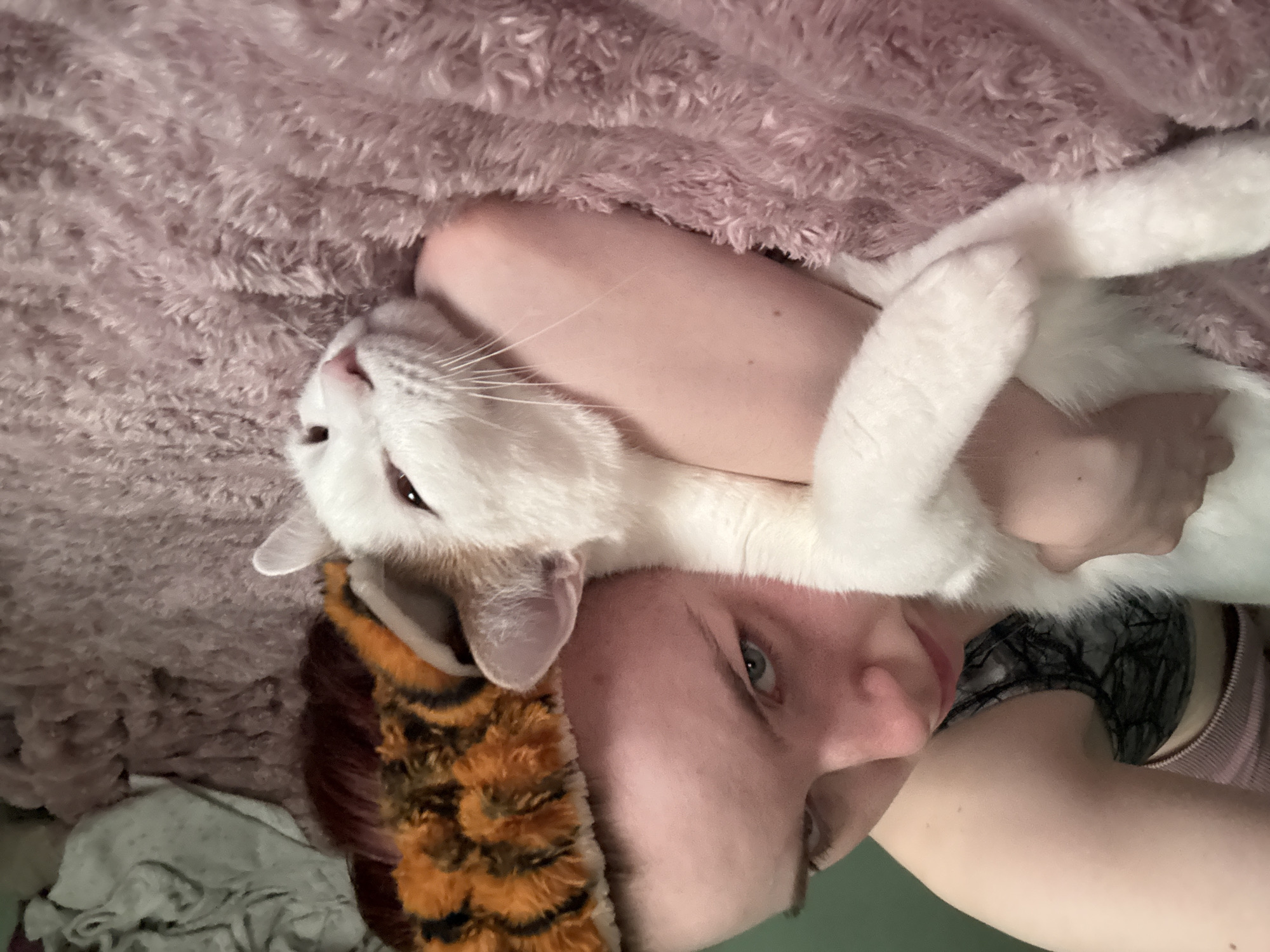 A person with red hair and a tiger-striped headband is lying down and holding a white cat with brown markings. The person's face is partially obscured by the cat, and they appear to be looking at the camera. The cat is resting its head on the person's arm and looks relaxed. They are both on a pink, fluffy blanket. The background is blurred and shows a green wall and some crumpled fabric. The scene appears to be indoors.