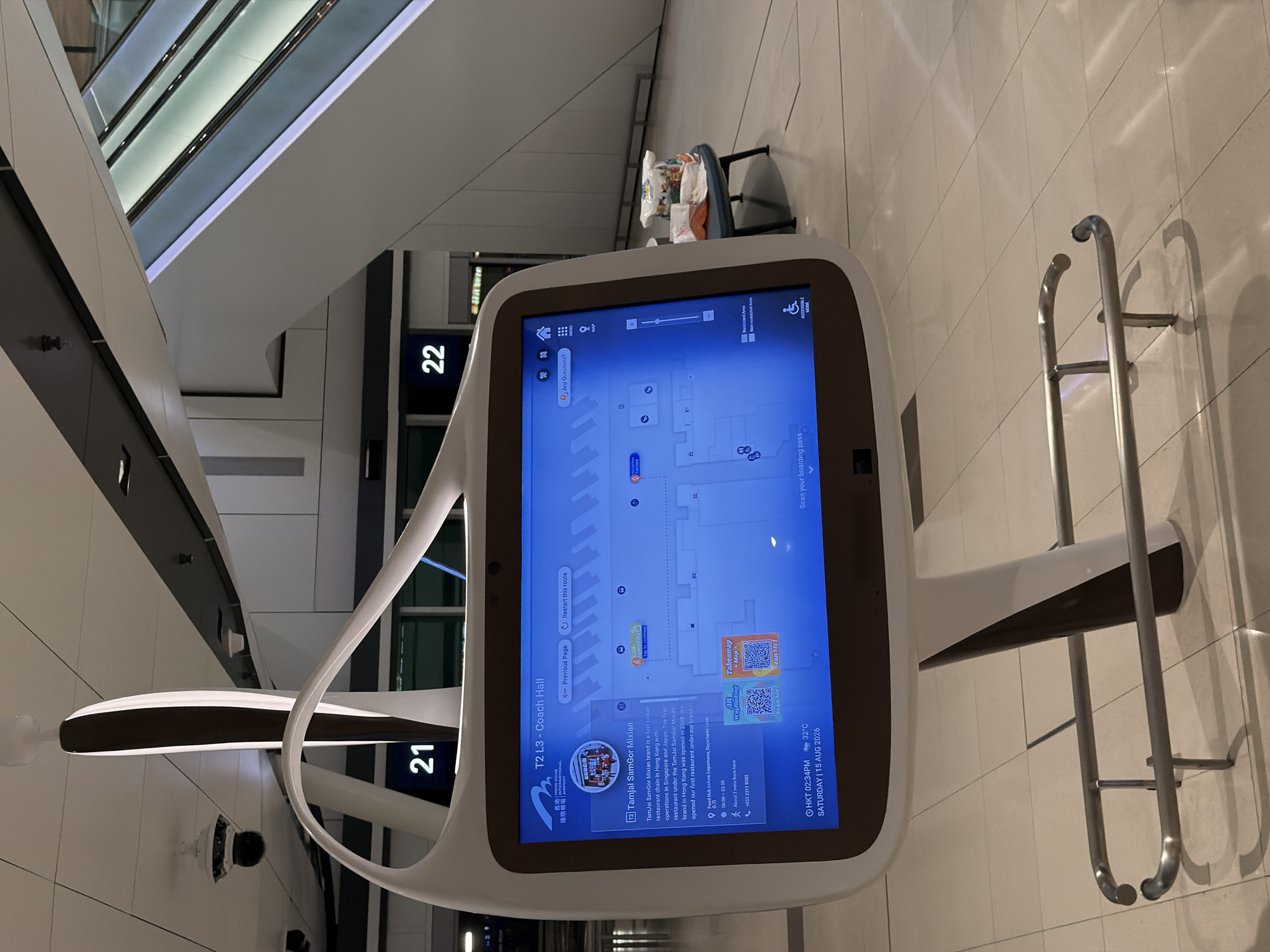 An interactive digital kiosk displays information about "T2 L3 - Coach Hall" at Hong Kong International Airport. The kiosk shows details about a restaurant called "TamJai SamGor Mixian," including its location, operating hours, and contact number. The screen also features a map of the area, options to scan a QR code for AR Wayfinding or Takeaway, and a button for "Accessible Mode." In the background, an escalator is visible, along with signs indicating "21" and "22." A small table with several bags of snacks sits to the right of the kiosk. The environment appears to be an indoor, modern airport terminal with tiled flooring and a high ceiling.