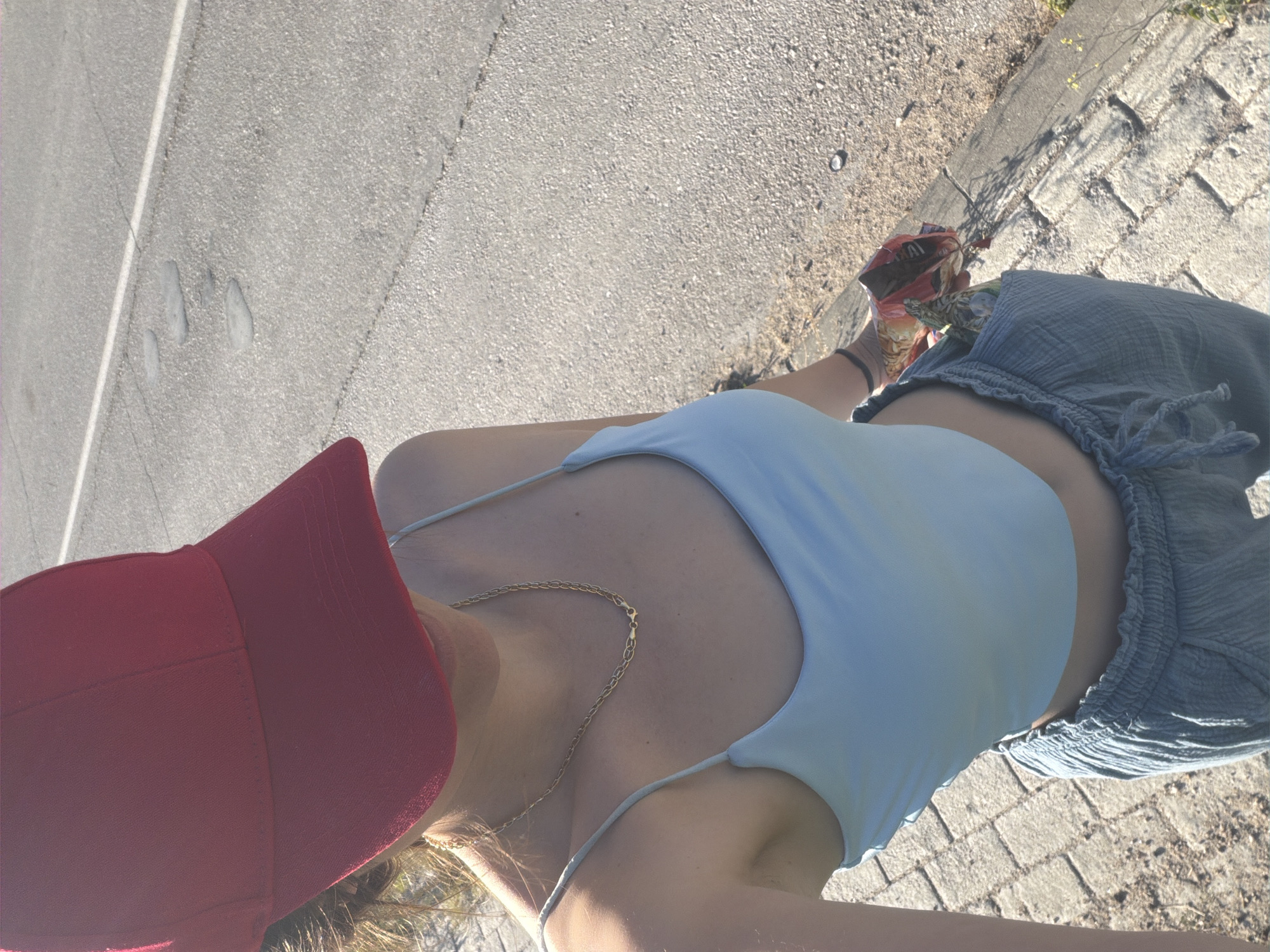 A woman wearing a light blue top, grey shorts, and a red baseball cap stands on a brick and concrete sidewalk adjacent to an asphalt road. She holds a bag of chips and wears a gold chain. The location appears to be an outdoor public space in Lesichovo, Bulgaria.