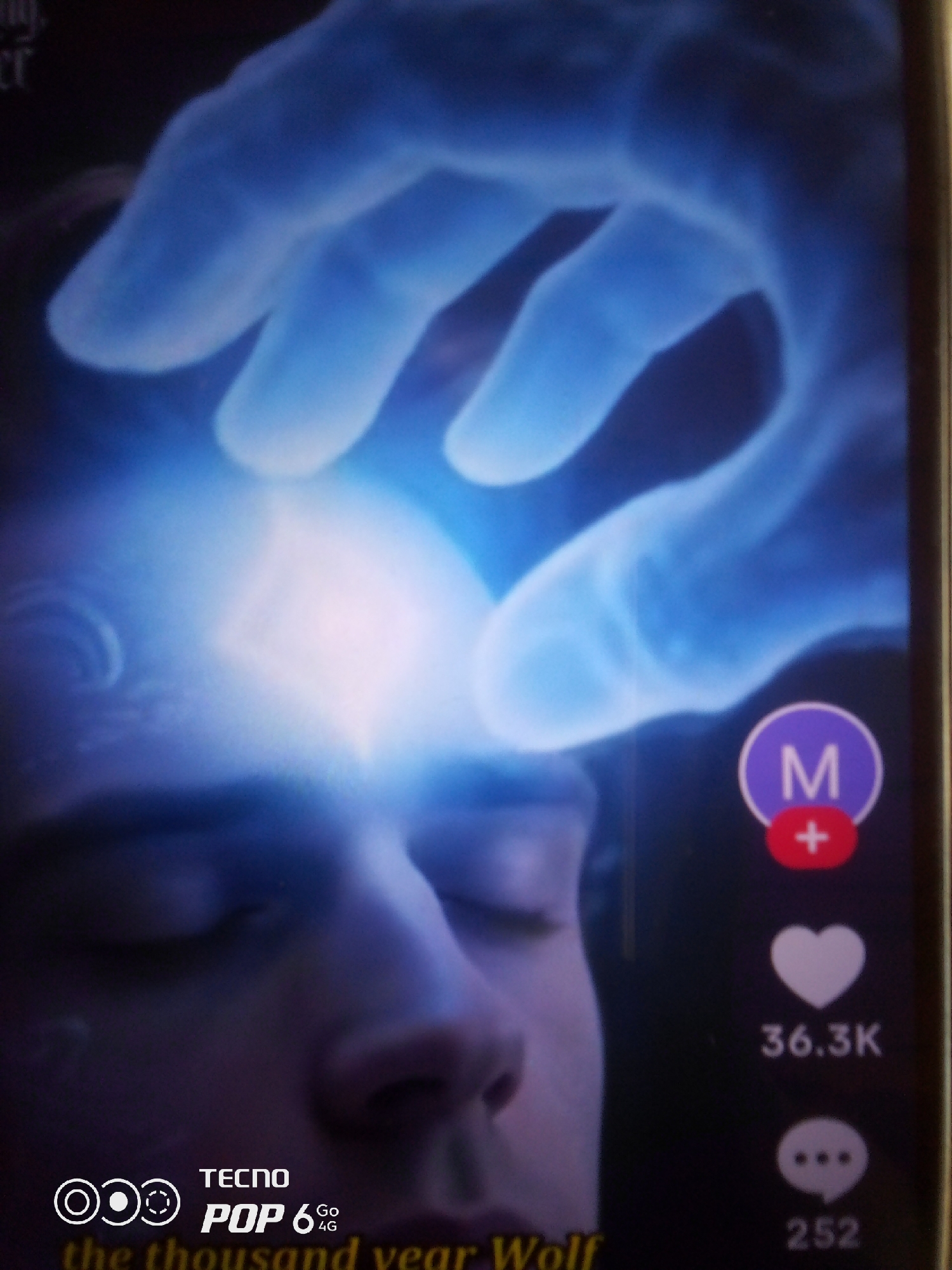 The image shows a close-up of a person's face, with their eyes closed and a bright, ethereal light emanating from their forehead. A large, translucent blue hand appears to be reaching down and touching the light. On the right side of the image, there are social media interaction icons: a purple circle with a white 'M' and a red plus sign, a white heart icon with the number "36.3K" below it, and a white speech bubble icon with the number "252" below it. At the bottom left, there is a logo that appears to say "TECNO POP 6 Go 4G" and some text that reads "the thousand year Wolf". The image is likely a screenshot from a video or social media post. There is no visual information in the image that would allow for the determination of a specific location within Howayi, Nigeria.