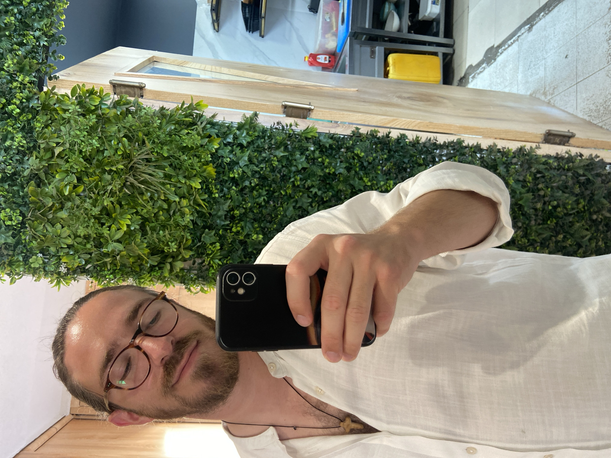 A selfie shows a bearded man in a white shirt and glasses, holding a smartphone. The background features a wall covered in artificial greenery. Above the greenery is a light wood structure, potentially a frame or part of a building. Further in the background, blurred objects suggest an interior space with shelving and various containers, some brightly colored (red and yellow). This could be in Petrovac centar, Montenegro given user specified location.