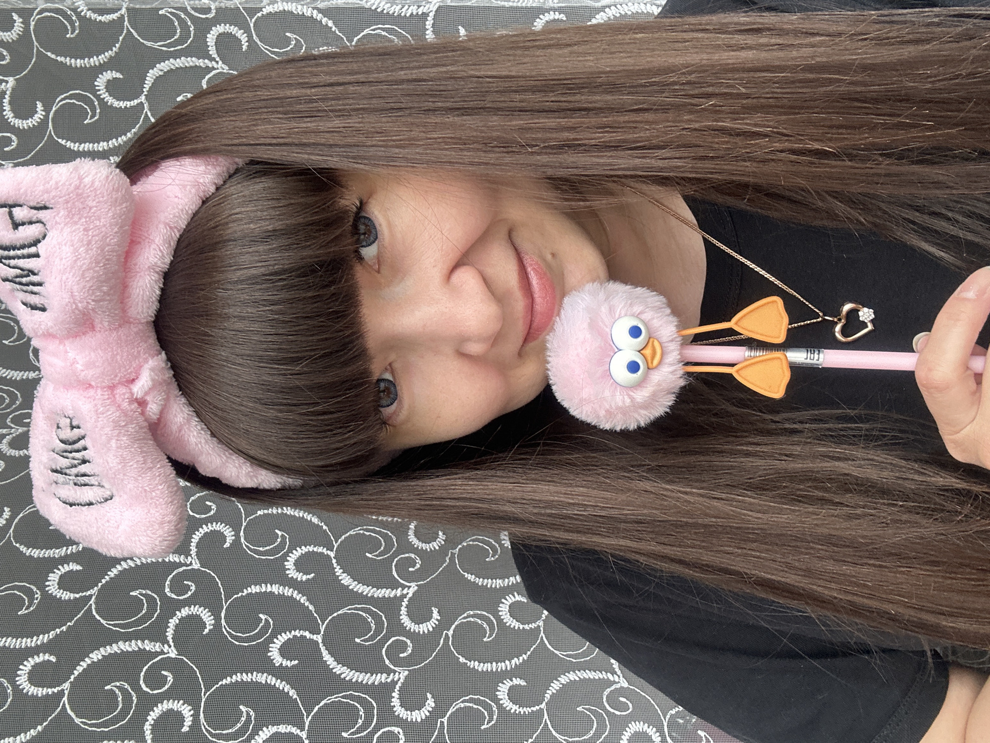 A medium shot shows a woman indoors, with dark brown hair and bangs, wearing a pink bow headband with writing on it. She is wearing a black top and a gold necklace with a heart-shaped pendant. She is holding a pink pen with a fuzzy, pink chick decoration with large blue eyes and orange feet. The background is a gray surface with a white floral pattern. The photograph was taken in the city of Chaikovsky, Russia.