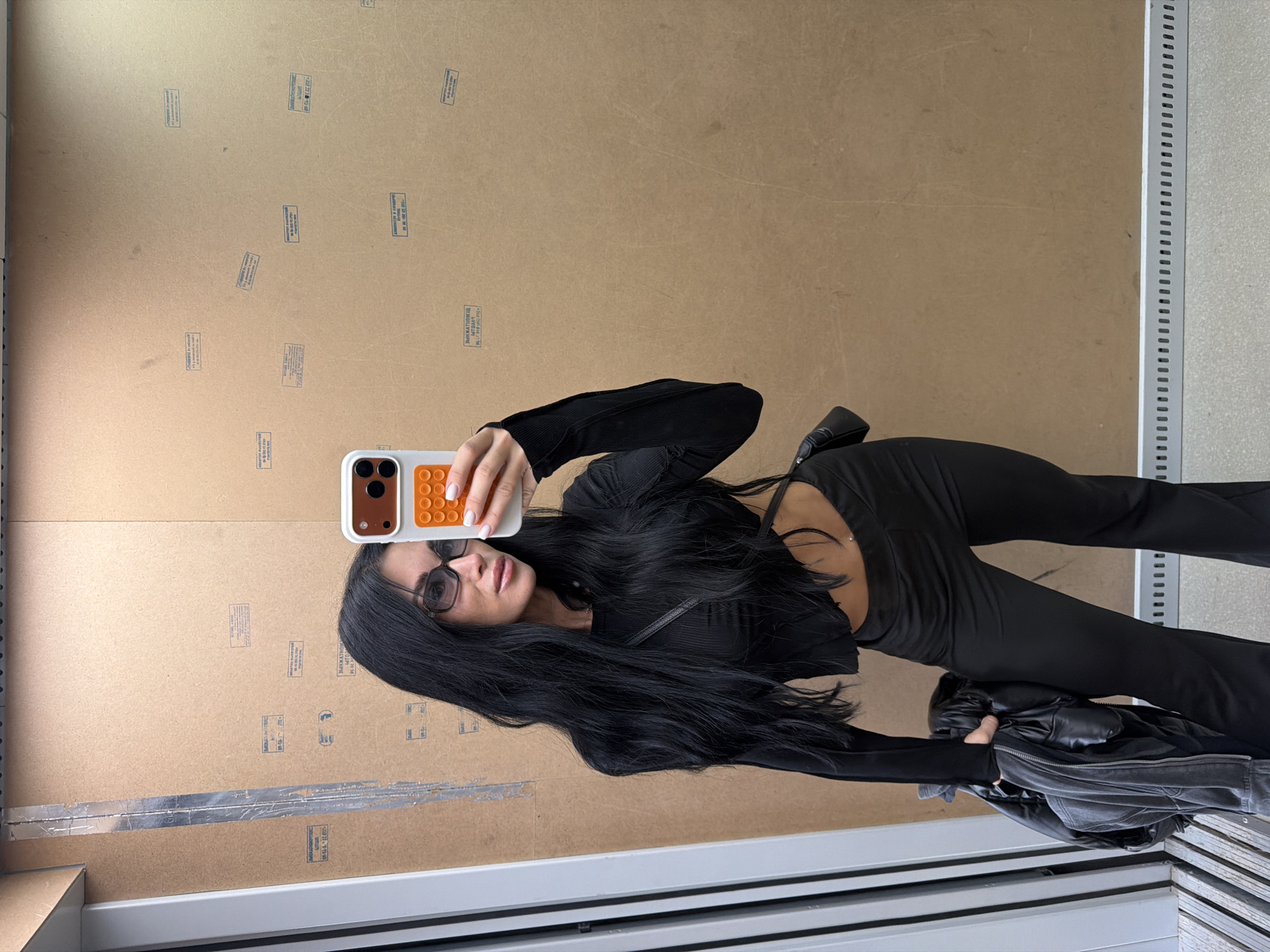 A woman with long, dark hair, wearing a black, long-sleeved, cropped top and black, flared pants, stands in front of a wall covered in various stamps and labels. She is holding a black, puffer jacket in her left hand and a smartphone in her right hand, taking a selfie. She wears dark sunglasses and a small black purse is slung across her chest. The environment appears to be an industrial or storage area, with a metallic vertical beam to the left and a grey tiled floor with a ventilation grate at the bottom. The wall features numerous stamped markings, some with Cyrillic text, suggesting it might be a shipping or manufacturing facility. The lighting is adequate, casting shadows and highlighting the textures of the wall and the woman's clothing. The image is taken indoors. There is no indication of the specific location within the city of Brest, Belarus, beyond what can be inferred from the general environment.