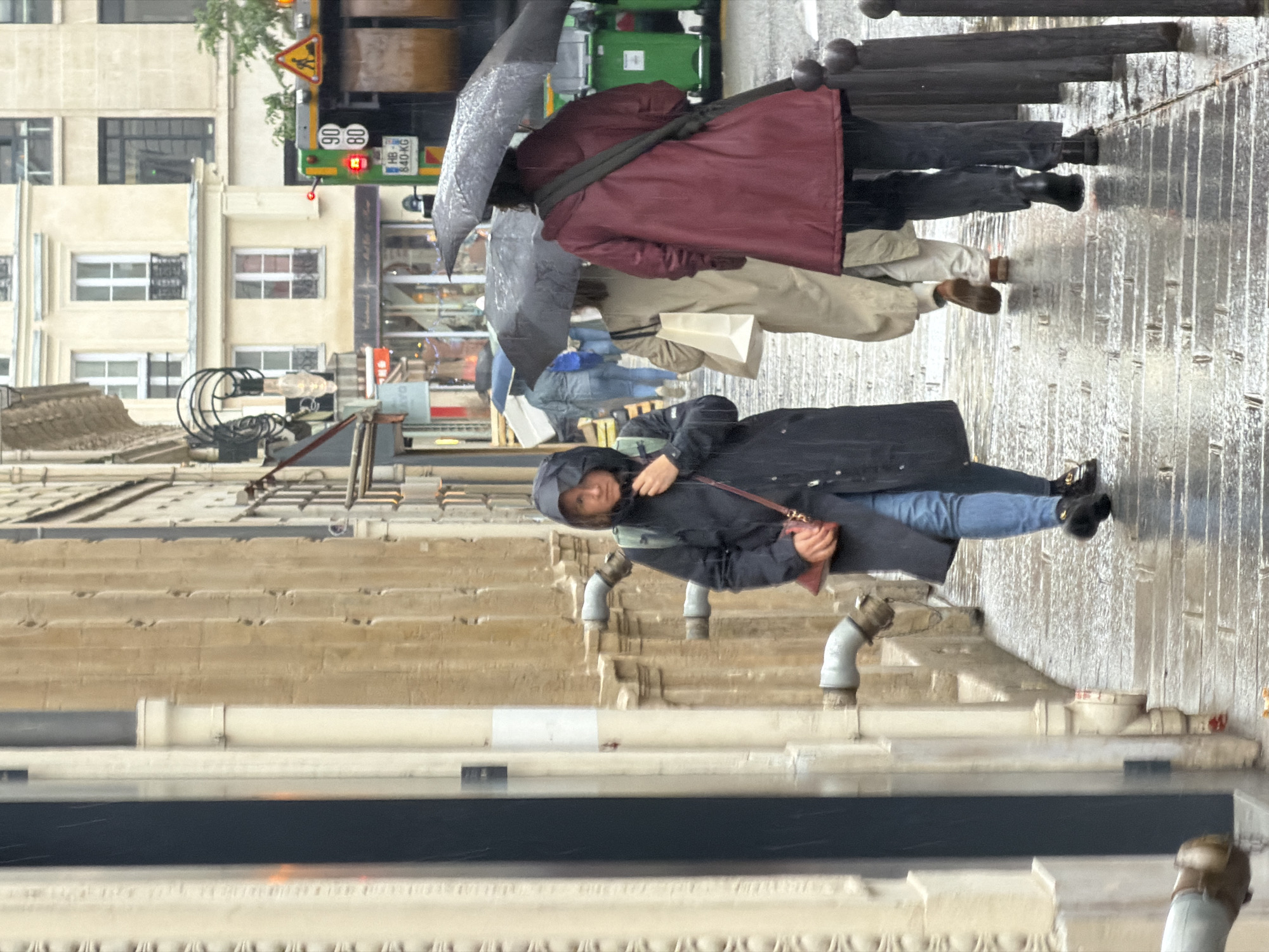 People walk down a wet sidewalk on a rainy day in Paris, France. A woman wearing a dark raincoat and jeans walks in the foreground. People in the background use umbrellas. A sign indicating a construction zone is visible. A building is in the background.