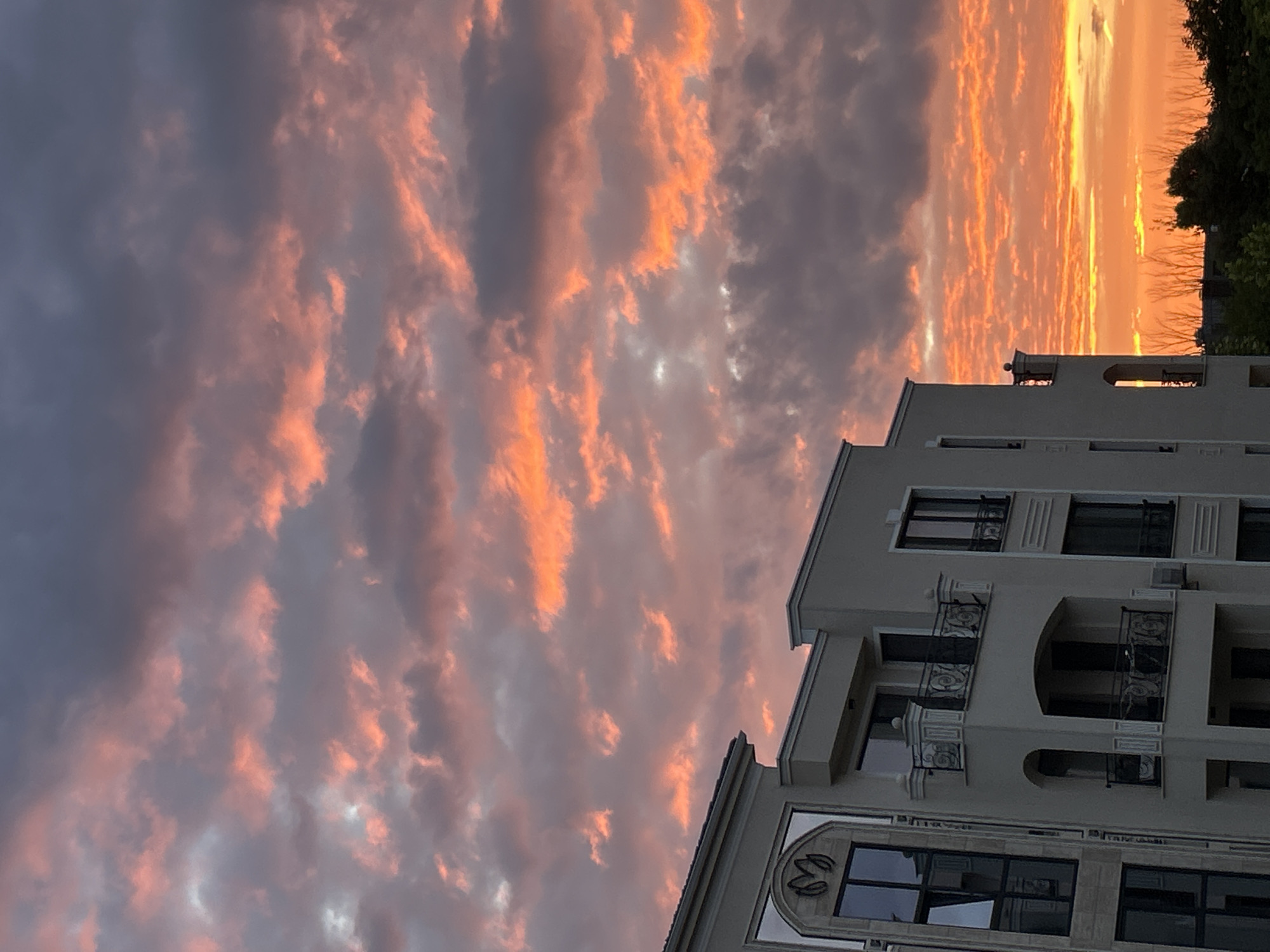 The image shows a building in Plovdiv, Bulgaria during a sunset. The sky is filled with dramatic clouds painted in hues of orange, pink, and grey. The building is modern with multiple floors and balconies, and is situated against the backdrop of the vibrant sky. Some trees are visible in the lower right corner.
