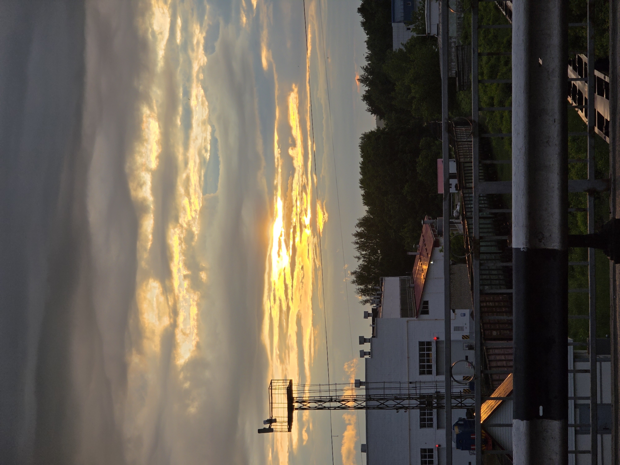 The image captures a dramatic sunset or sunrise over an industrial or urban landscape in Chaikovsky, Russia, taken from an elevated perspective, likely a bridge or overpass. The sky dominates the upper two-thirds of the frame, showcasing a striking display of clouds illuminated in vibrant golden and orange hues by the hidden sun. The sun's intense glow is visible peeking through thick, elongated clouds, casting brilliant streaks of light across the sky and reflecting on surfaces below.
Along the horizon, a dense line of dark green trees is silhouetted, particularly on the right side of the image. Below the treeline, several buildings with light-colored walls and some red roofs are visible. One prominent white building in the mid-ground features multiple windows, dark ventilation units on its roof, and a small red light on its facade. To its left stands a tall, slender metal lattice tower with a platform at its top, adding an industrial element to the scene. The golden light of the sun reflects noticeably on the red roof of one building and parts of the foreground structure.
The foreground on the right is occupied by a robust metal fence or railing, with vertical and horizontal bars in shades of grey and black, suggesting the viewpoint is from an elevated structure. No human activity or visible text is present. The overall atmosphere is serene yet dramatic, emphasizing the natural beauty of the sky contrasting with the man-made environment.