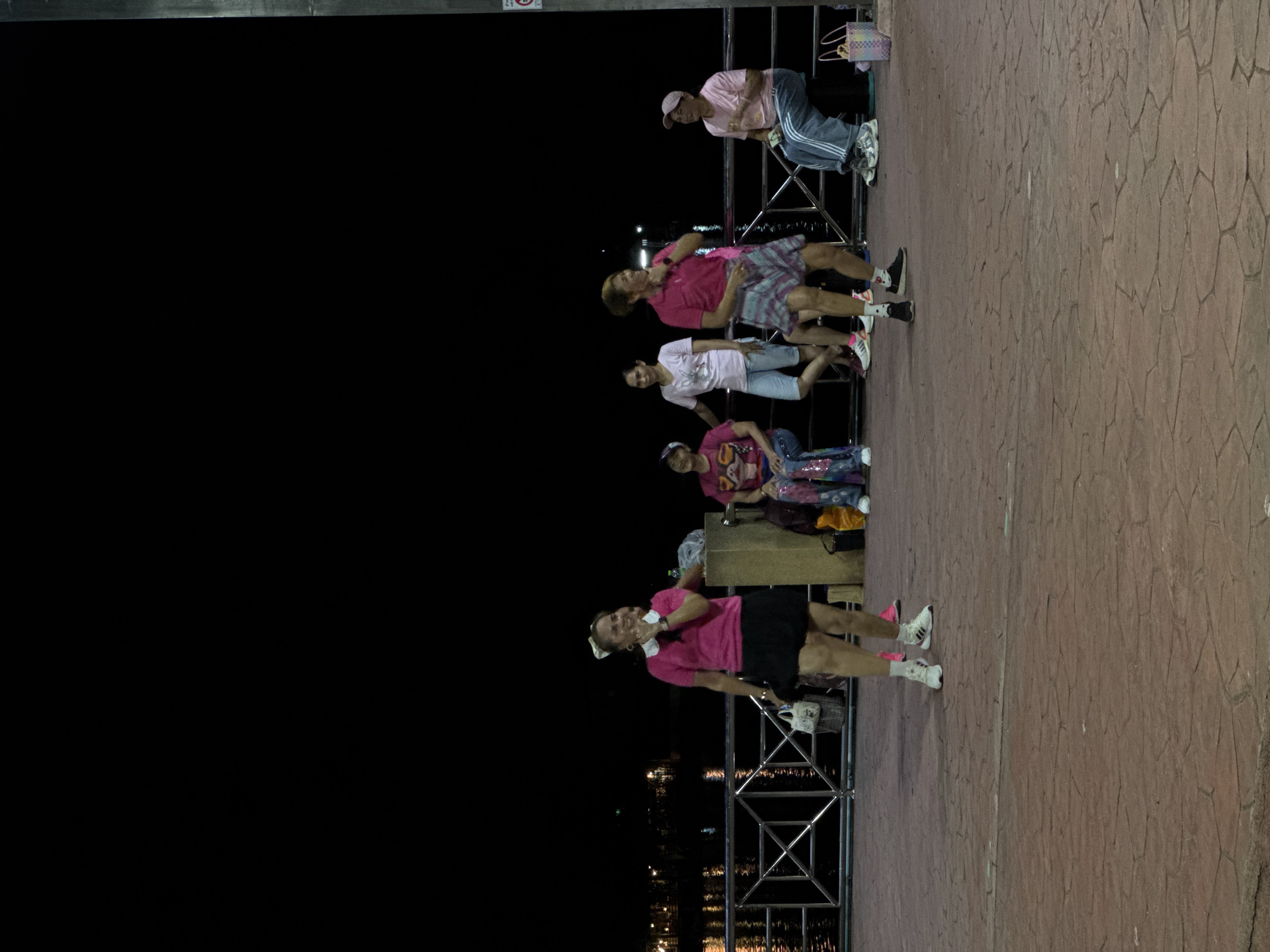 The image depicts an outdoor nighttime scene, likely in a public area with a paved ground and a railing overlooking a body of water with distant lights. Several people, mostly women, are present.
In the foreground, a woman in a pink short-sleeved shirt and a black skirt is dancing or posing, with her left hand raised. She has a white bow in her hair and is wearing white athletic shoes.
To her right, a woman in a pink shirt and a plaid skirt is also in motion, with her right hand near her face. She is wearing black athletic shoes.
Further to the right, another woman in a pink t-shirt and a cap is sitting on a dark stool, holding a bag. She is wearing grey sweatpants and athletic shoes.
Behind the dancers, other individuals are visible. One woman in a pink t-shirt and sparkly pants is sitting. Another woman in a white t-shirt and light blue shorts is standing and leaning on the railing. A woman in a pink t-shirt with a graphic design on it is also seated.
Objects in the scene include a wooden post, various bags, buckets, and a pink and white woven basket. The railing is made of metal with a geometric pattern. The paved ground has a textured, stone-like appearance. The background shows the dark expanse of water and the illuminated skyline of Surat Thani City Municipality, Thailand.