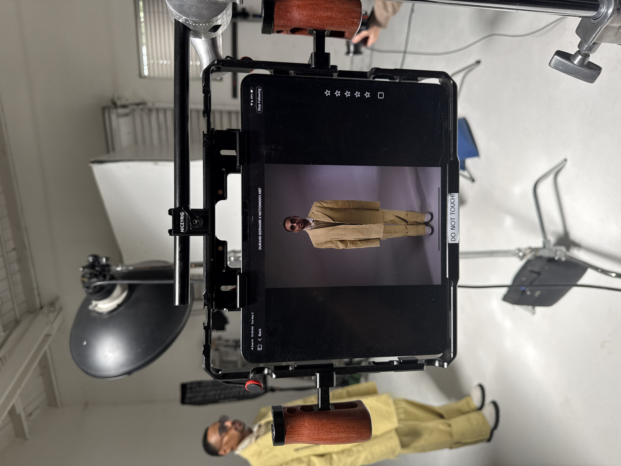 The image captures a behind-the-scenes look at a photoshoot or filming session. A tablet mounted on a rig displays a photo of a person wearing sunglasses, a light brown suit, and a tie. The tablet’s screen shows information such as the photo’s file name "DURAND BERNARR X NOTION0001.NEF" and the date "Tue May 5". The screen also includes a "DO NOT TOUCH!" sticker. The rig holding the tablet is attached to lighting equipment, including a black reflector, and a metal stand with a wooden handle. The environment appears to be a studio with a white background. There is no evidence of location in Los Angeles, United States of America.