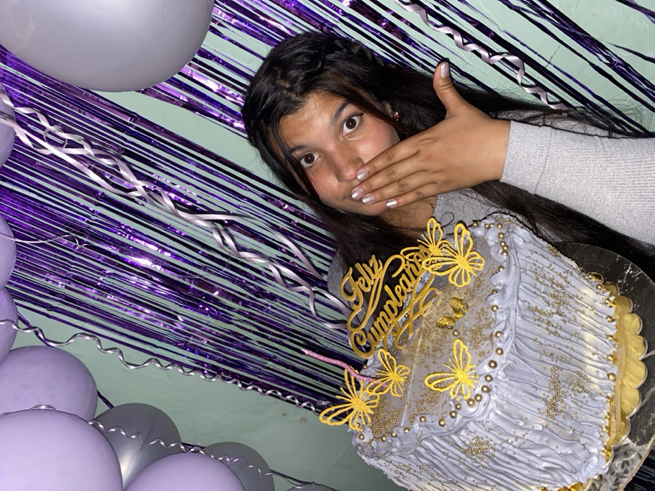 A young woman is posing with a celebratory birthday cake. She has dark hair and a grey sweater. Her hand is covering her mouth, and she’s looking toward the viewer. The birthday cake is light purple with gold embellishments and a “Feliz Cumpleaños” (Happy Birthday) topper. The background consists of purple and silver streamers and light purple and silver balloons.
