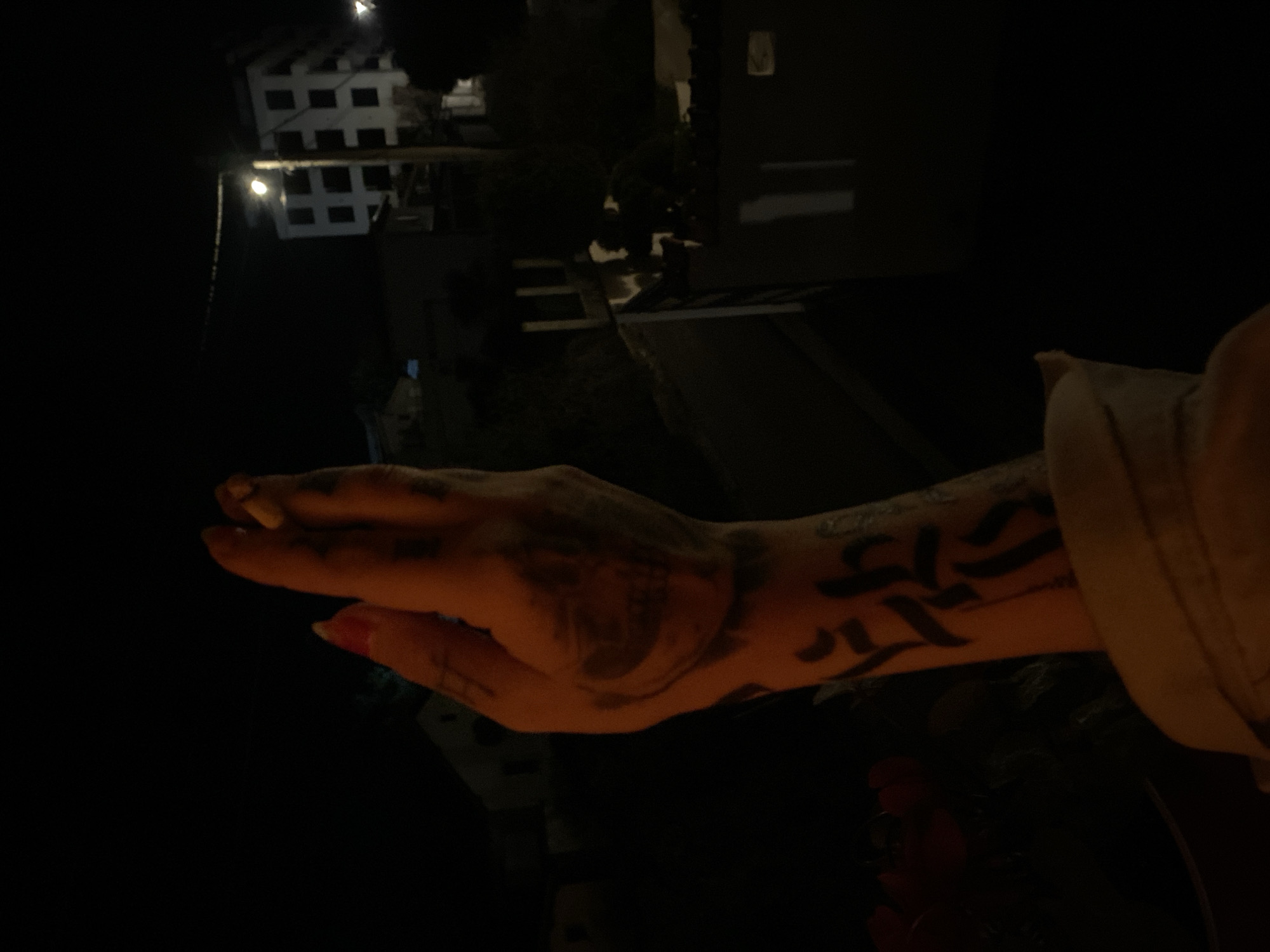 A person's tattooed hand holds a lit cigarette. The background suggests an urban environment at night. A multi-story building with illuminated windows is visible. A walkway or road is also present. The location is Unknown, Unknown.