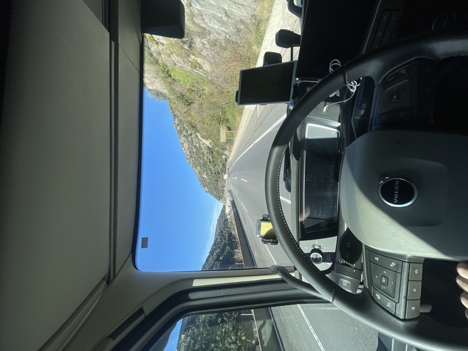 The image presents a first-person view from the driver's seat of a Volvo heavy goods vehicle. A human hand is visible on the lower-left section of the multi-function steering wheel, which bears the "VOLVO" emblem. The digital instrument cluster displays "70", indicating the vehicle's current speed. On the left side of the dashboard, a yellow and black mobile device mount is attached. To the right, a dark-screened tablet is secured in a black mount, with white charging cables visible beneath it.
The vehicle is in motion on a multi-lane highway, characterized by asphalt surfacing and multiple lanes of travel. The highway is bordered on both sides by mountainous terrain featuring steep, rocky slopes partially covered with vegetation. On the right-hand shoulder, a brown road sign is discernible, displaying the text "MUSEU MEMORIAL L'EXILI", which accurately locates the scene in the vicinity of La Jonquera, Spain. The sky is clear and blue, indicating bright daylight conditions. Several other vehicles are present further down the highway in the distance.