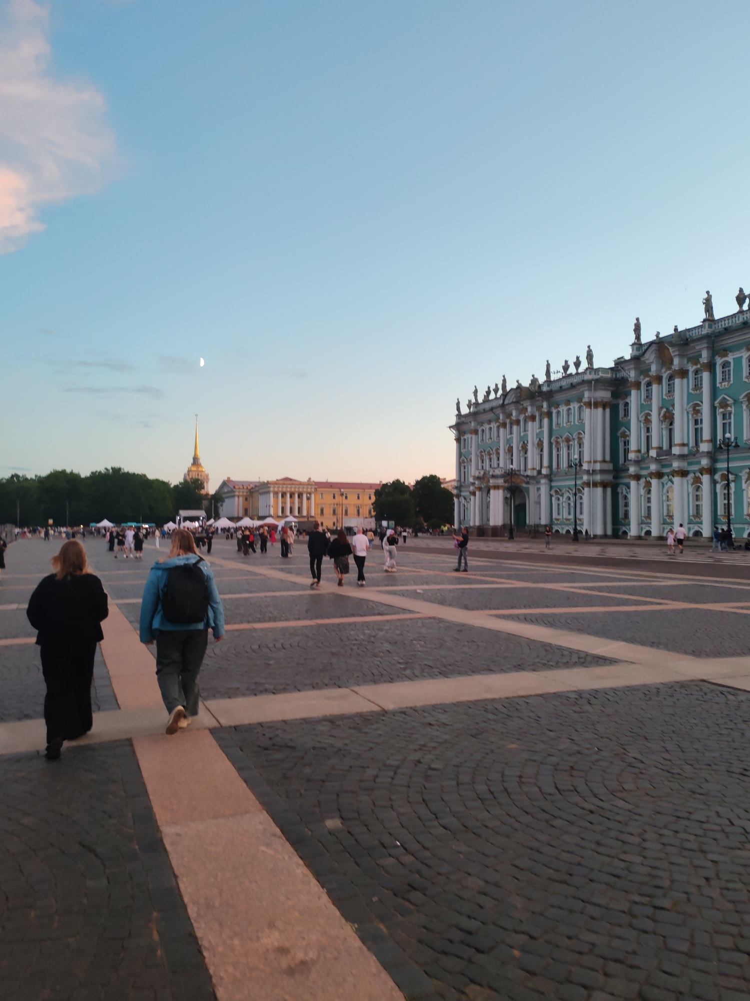 This image depicts Palace Square in Saint Petersburg, Russia, at dusk. The iconic Winter Palace, with its turquoise facade and ornate white columns, dominates the right side of the frame. In the background, across the vast cobblestone square, the Admiralty Building's golden spire pierces the pale blue sky, which is illuminated by soft pastel clouds. A sliver of a moon is visible in the upper left quadrant.
Several people are present in the square, mostly walking or standing in small groups. Two women are in the foreground, walking away from the camera towards the left. The woman on the left is wearing a black dress, and the woman on the right has on a blue jacket and dark pants, carrying a backpack. Other individuals and groups are scattered throughout the square, suggesting a public gathering or a popular tourist spot. The ground is paved with large cobblestones, with lighter colored rectangular stones marking pathways. The overall atmosphere is calm and serene, characteristic of a late evening in a grand European city.