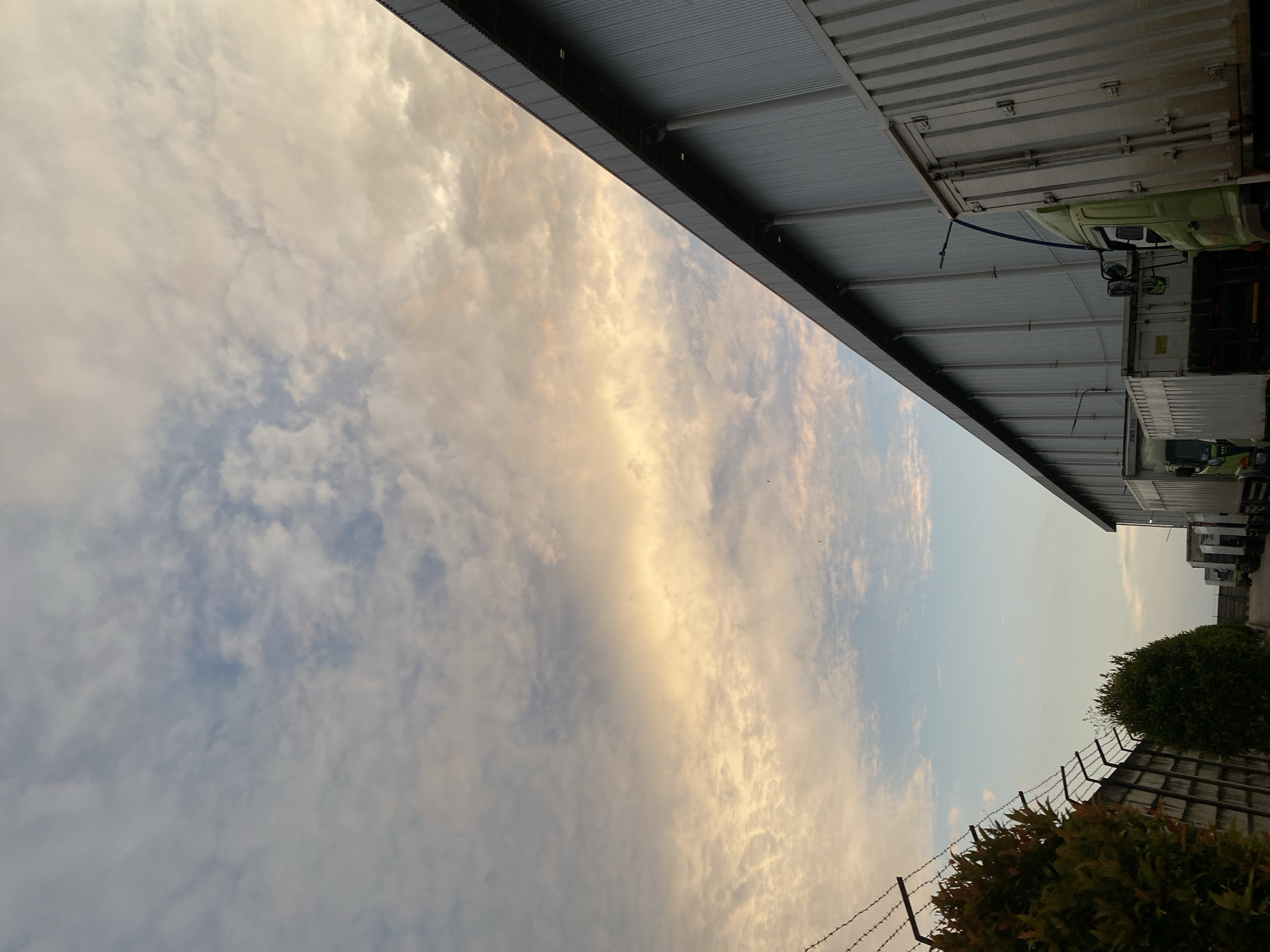 This photograph captures a twilight scene at a logistics facility in Gambut Barat, Indonesia. The sky above is filled with a mix of pale blue and warm, muted orange and yellow clouds, indicative of either sunrise or sunset. In the foreground, a corrugated metal building, likely a warehouse or factory, dominates the right side of the frame, its long facade stretching towards the horizon. A line of parked trucks is visible along the side of the building, their metallic cargo containers reflecting the ambient light. To the left, a fence topped with barbed wire separates the facility from a row of dense, dark green foliage, possibly trees or large bushes. The ground appears to be a dirt or gravel surface. The overall impression is one of industrial activity paused during the transitional light of day. There are no people visible in the frame.