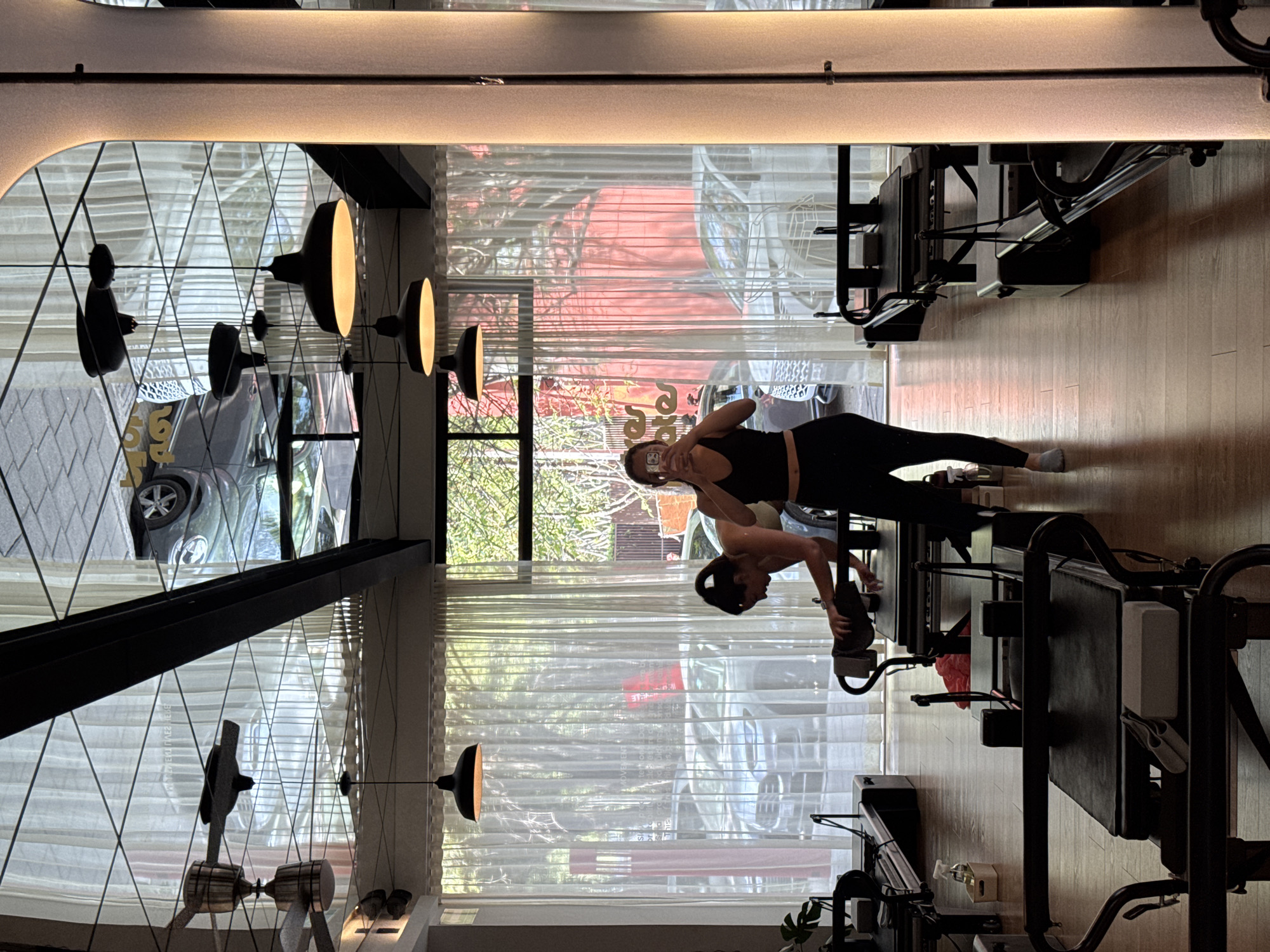 The image depicts an indoor scene, likely a Pilates or fitness studio, characterized by modern design and ample natural light. The room features light wooden flooring, large windows, and sheer white curtains that partially obscure the view outside.
The main subject is a woman, dressed in a black athletic top and black leggings, standing near a Pilates reformer. She is holding a smartphone to take a selfie, with her reflection clearly visible in the window or mirror. Her hair is pulled back, and she is wearing light-colored socks.
Several black Pilates reformers are visible in the foreground and background, suggesting an active fitness environment. Modern, minimalist pendant light fixtures with warm-toned interiors hang from the ceiling, adding to the contemporary aesthetic.
Notable details include reflections in the large windows, showing the interior of the studio and parts of the outdoor environment. Outside, a dark-colored car, appearing to be a Volkswagen Beetle, is distinctly reflected. Trees and possibly other buildings are also faintly visible through the curtains.
Visible text, albeit heavily distorted and mirrored due to reflections on the sheer curtains, includes words such as "MOVERS" and "STRETCHES" or "STRETCH", possibly part of a studio name or motto. The image is taken during daytime, evidenced by the bright natural light filtering through the windows. The overall atmosphere suggests a serene and functional fitness space.