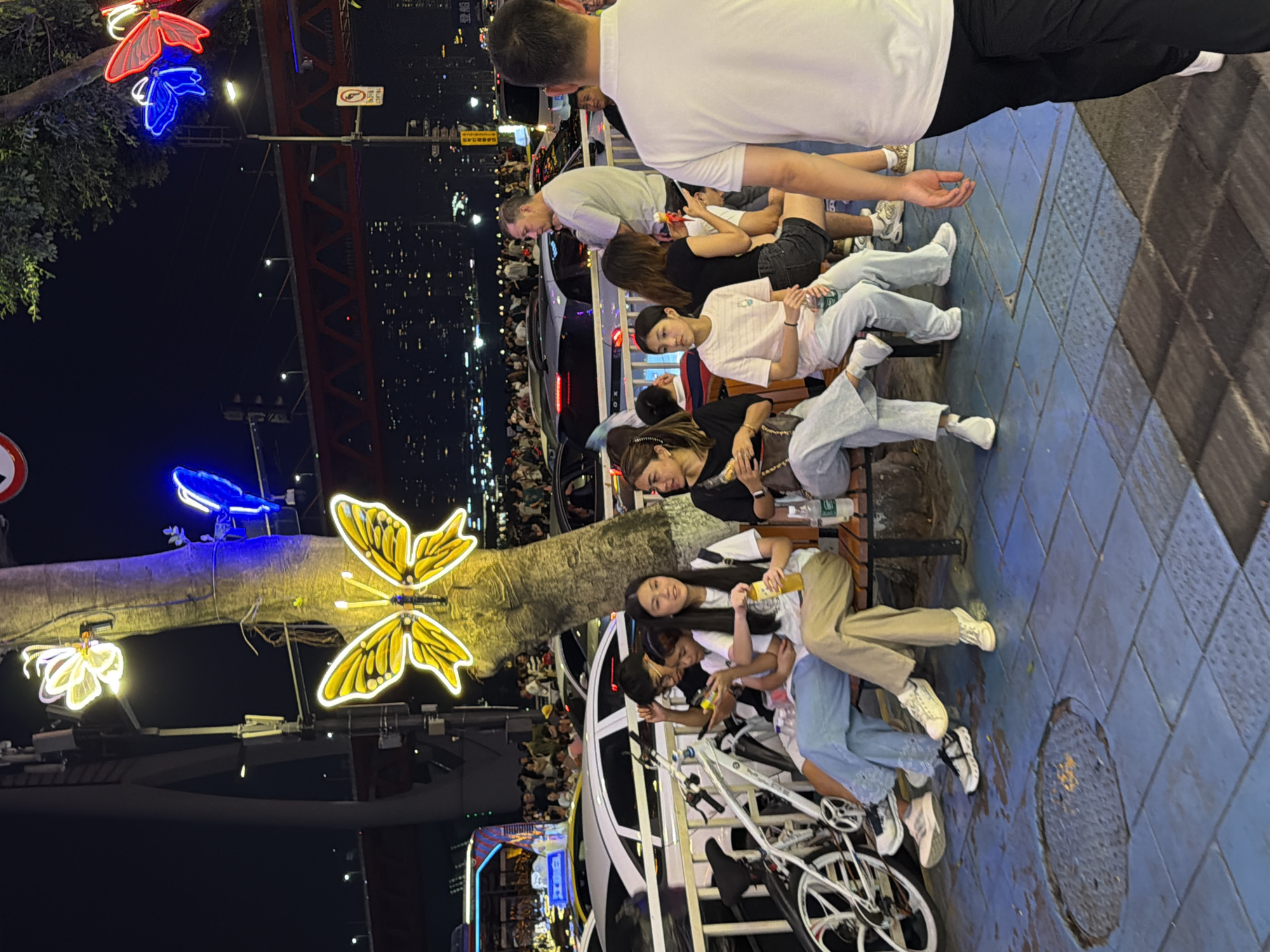 The image depicts a nighttime scene on a street in Yuzhong District, China. Several people are seated on a bench in the foreground, some holding drinks and interacting casually. A bicycle is parked nearby. In the background, a bustling street scene unfolds with numerous people, vehicles, and illuminated buildings. Decorative butterfly lights are prominent features, attached to a tree and possibly streetlights. The environment appears to be a pedestrian walkway or a promenade. The overall atmosphere suggests a lively urban setting during the evening.