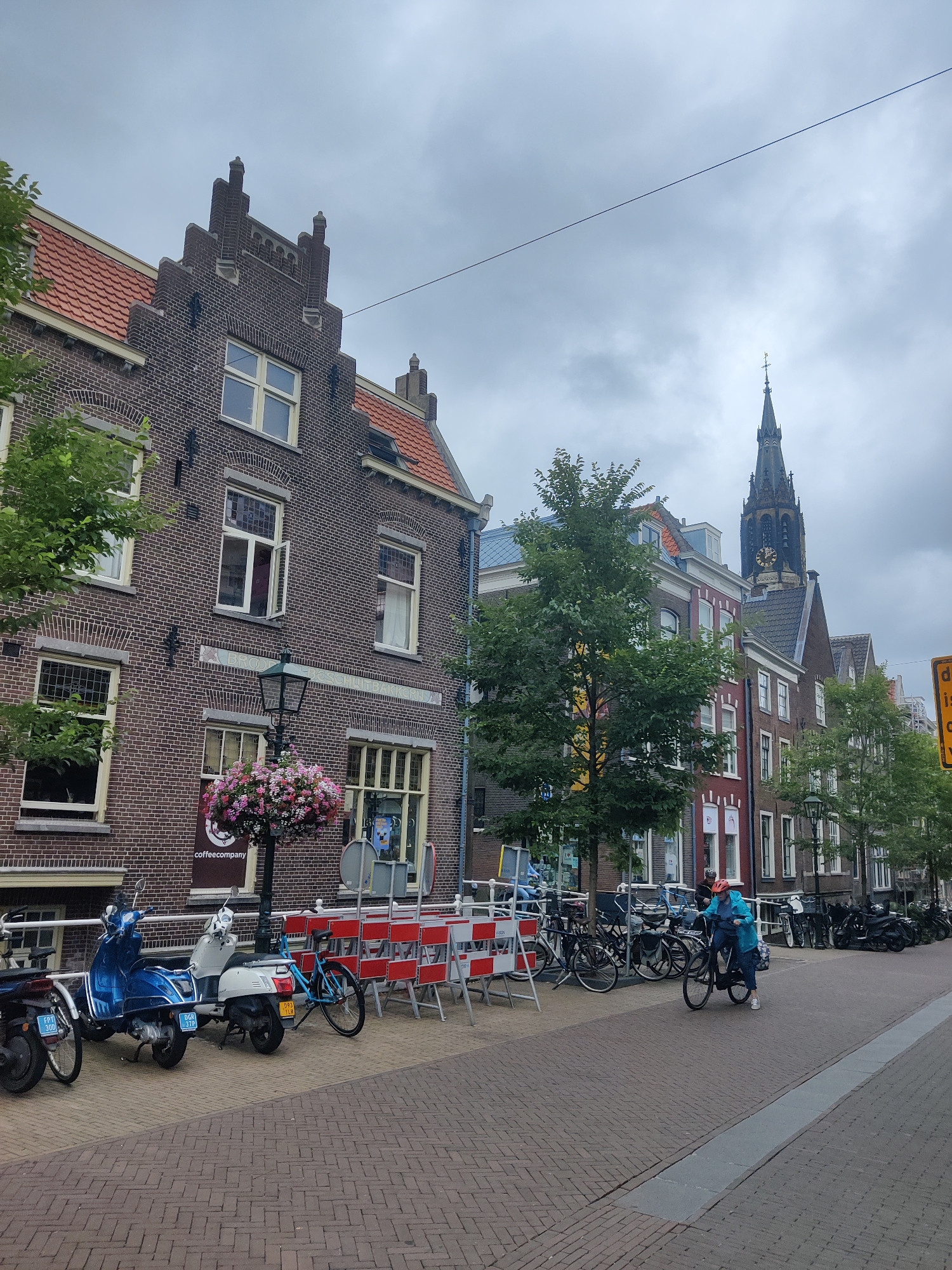 The image depicts a street scene in Delft, Netherlands. In the foreground, the brick-paved sidewalk is lined with parked scooters and bicycles. A blue scooter and a white scooter are parked next to each other, with license plates visible. Red and white barricades are set up along the sidewalk, suggesting ongoing construction or maintenance. Further down the street, a person wearing a blue jacket and an orange helmet is riding a bicycle.
The buildings lining the street are traditional Dutch architecture, made of brick with pitched tile roofs. One building features a prominent sign for "coffeecompany". In the background, the spire of a large church, likely the Nieuwe Kerk, rises above the rooftops, a distinctive landmark of Delft. The sky is overcast, contributing to a muted lighting condition. Green trees are interspersed along the street, adding a touch of nature to the urban environment. The overall scene conveys a typical day in a Dutch city, with a mix of historical architecture and modern modes of transportation.