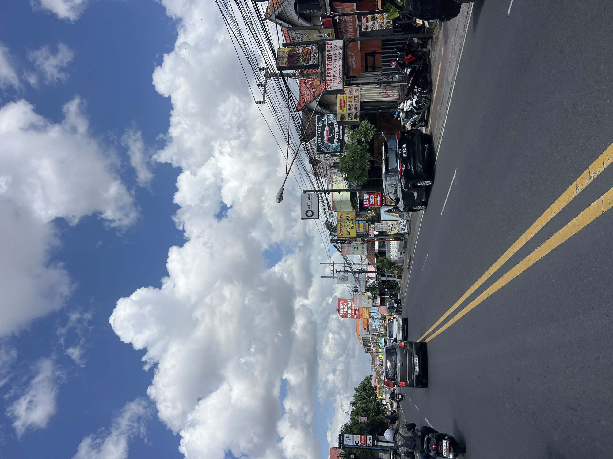 This is a wide-angle, eye-level shot of a busy street in Denpasar, Indonesia, on a bright, cloudy day. The road, with double yellow lines, is filled with traffic including cars, motorcycles, and a scooter. Buildings line the right side of the street, adorned with numerous signs, indicating shops, restaurants, and possibly services. These signs are primarily in Indonesian and feature vibrant colors and images. Prominent among them are advertisements for food items like "SATE BABI GULING" and "LUMPIA SEMARANG." A power pole with a dense network of wires stands tall, adding to the urban landscape. In the distance, more buildings and trees are visible, suggesting a sprawling cityscape. The sky is a brilliant blue, dotted with large, fluffy white clouds, creating a dramatic contrast with the bustling street below.