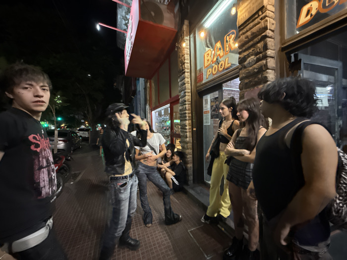 A group of people stand outside a "Bar Pool" establishment at night. On the left, a person with bangs wearing a black shirt faces the camera. Two others, one in a black jacket and the other in a white shirt, are in conversation. Three women stand near the bar entrance, and a person with a backpack stands slightly behind them. The scene is illuminated by the bar's lighting, and the street is visible with parked cars and trees in the background.