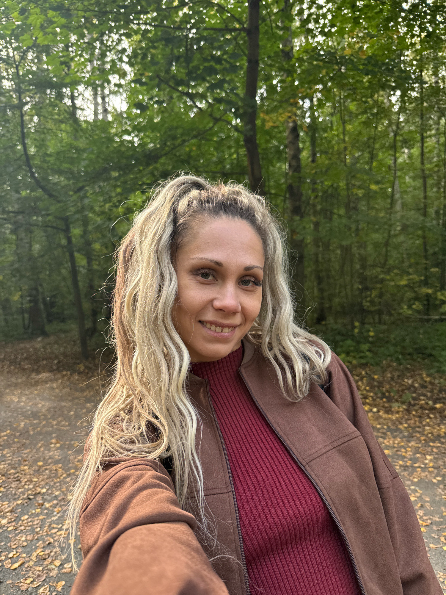 A woman is taking a selfie in a forest. She has blonde hair styled in a high ponytail and is smiling at the camera. She is wearing a burgundy turtleneck sweater and a brown jacket. The background shows a forest with green trees and fallen leaves on the ground. The location is in the city of Rusanovo, Russia.
