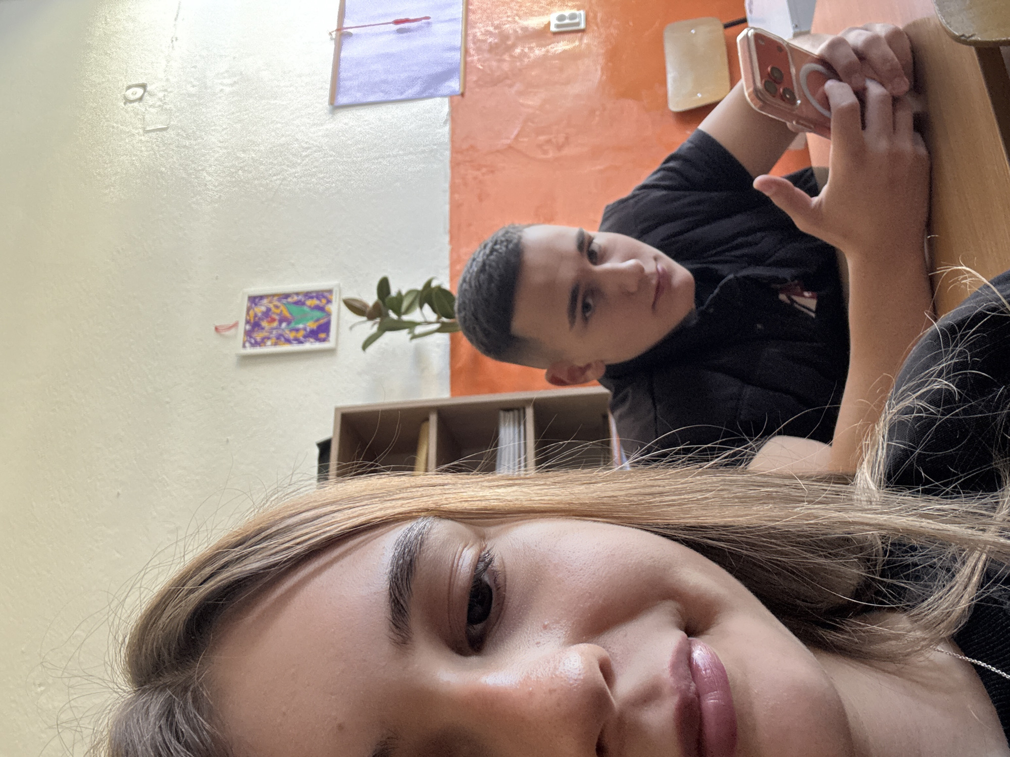 A selfie photograph shows two individuals, likely a young adult female and a young adult male, indoors. The female is in the foreground, her face close to the camera. She has long blonde hair and is smiling slightly. Behind her and slightly above, the male is holding a phone. He has short, dark hair and is looking at the camera. They are both wearing dark clothing. The background includes a wall with a color scheme of white and orange. A picture and a purple sheet are hanging on the wall. Additionally, there are some items and furniture visible in the background, including a plant. The image provides no explicit information to confirm the location is Panagyurishte, Bulgaria. The photograph is taken at an angle, giving a tilted perspective of the scene.