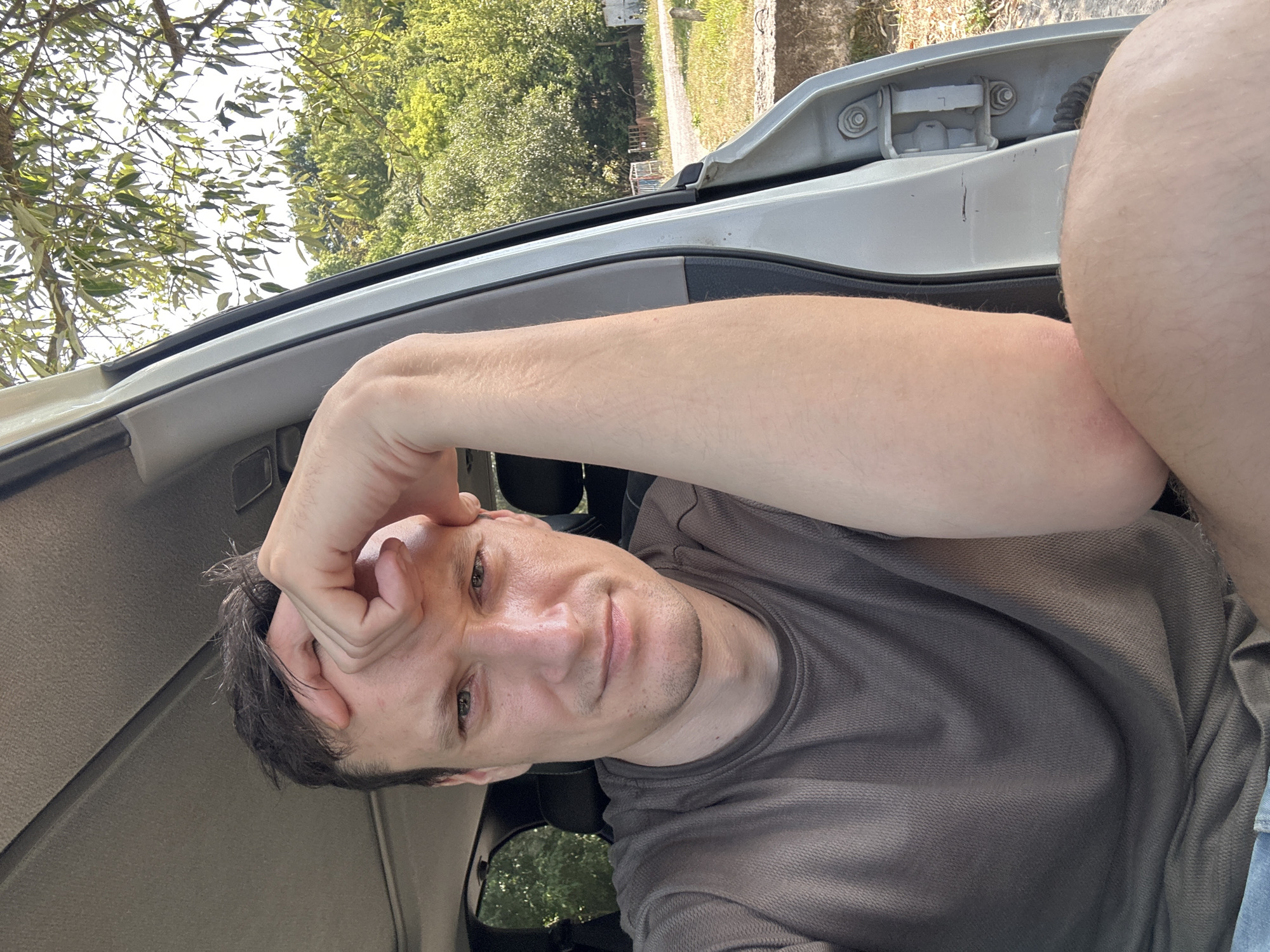 In Makiv, Ukraine, a man is captured in a selfie from the chest up, seated inside a vehicle with the door open. He is resting his forehead on his hand, which is placed over his brow. His gaze is directed towards the camera. The background reveals a sun-drenched outdoor environment with lush green trees and foliage. The interior of the vehicle is visible, including the headliner and part of the car door frame. The lighting suggests it is daytime, likely during the warmer months.