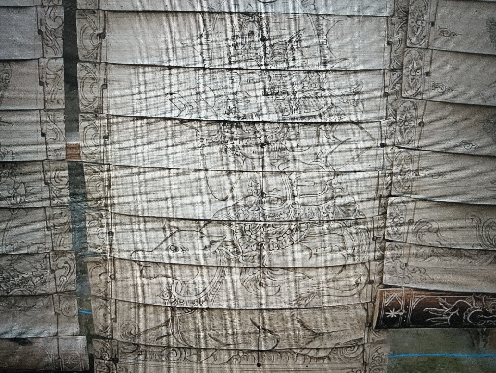 The image depicts a detailed drawing on a segmented material, possibly wood or canvas, forming a vertical panel. The drawing features a central figure, likely a deity or royalty, adorned with elaborate headwear and intricate clothing. Below this figure, a stylized animal, resembling a cow or bull, is depicted. The artwork exhibits traditional motifs and patterns, common in Indonesian art, especially from areas like North Sumatra, where the city of Medan is located. The material itself appears to be aged and textured, with visible lines and divisions between segments, suggesting it could be part of a larger decorative screen or mural. The overall impression is one of cultural artistry and historical significance.