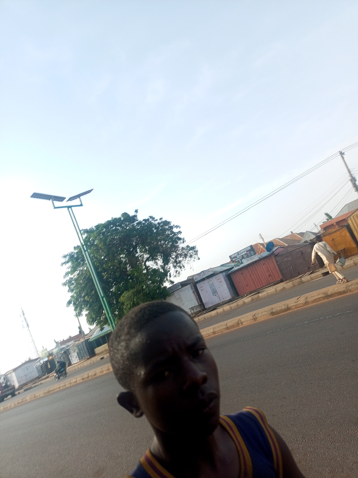 The image appears to be taken on a street in Tempure, Nigeria, on a sunny day. The foreground shows a young male facing the camera. He is wearing a dark blue and yellow tank top. The background shows a row of small shops and shipping containers. A man is walking along the road in front of the shops. A solar powered street light stands tall with a nearby tree. A motorcycle is further down the street.